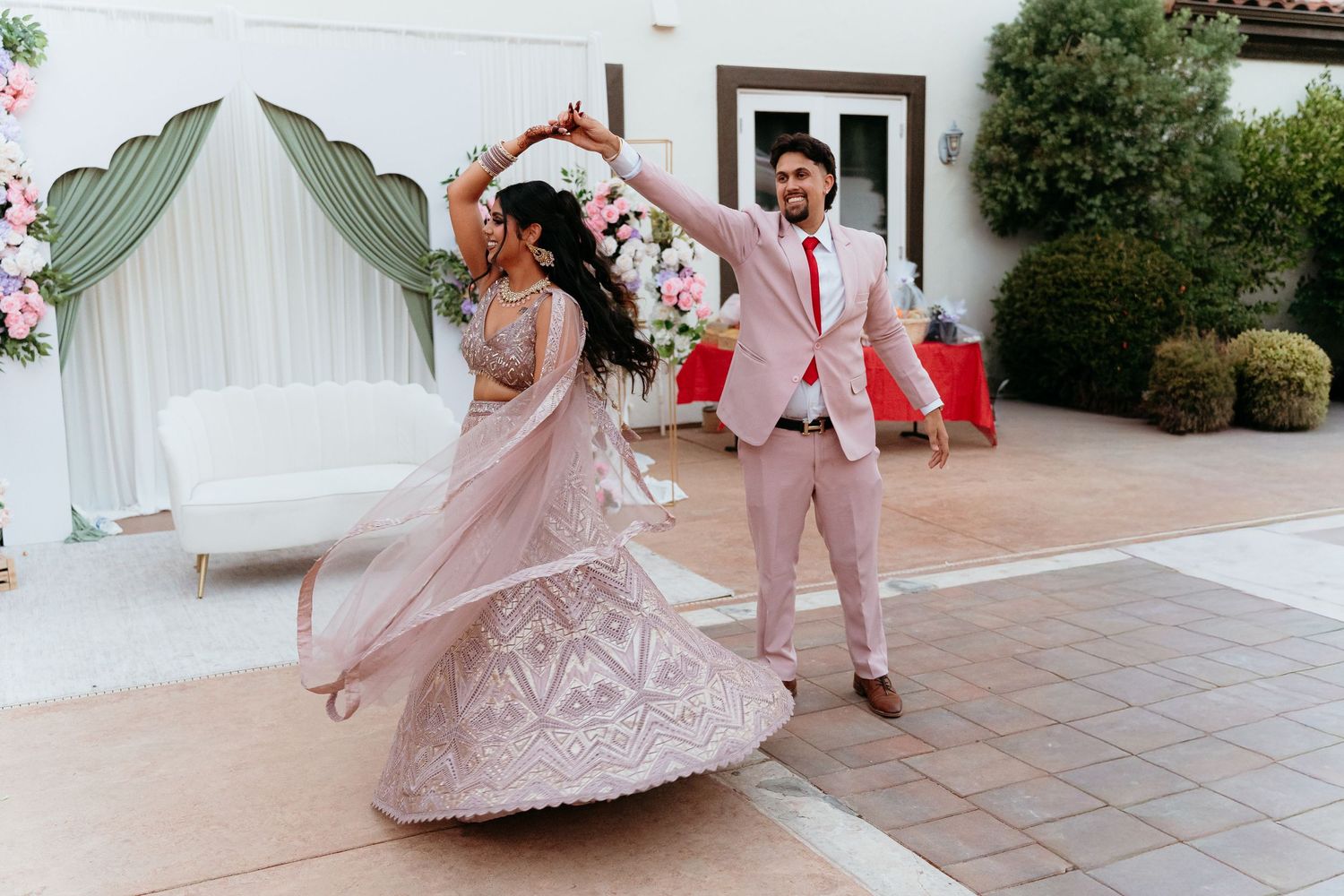Couple dancing at Indian wedding, bride in pink lehenga twirling, groom in blush suit, floral backdrop outdoors.