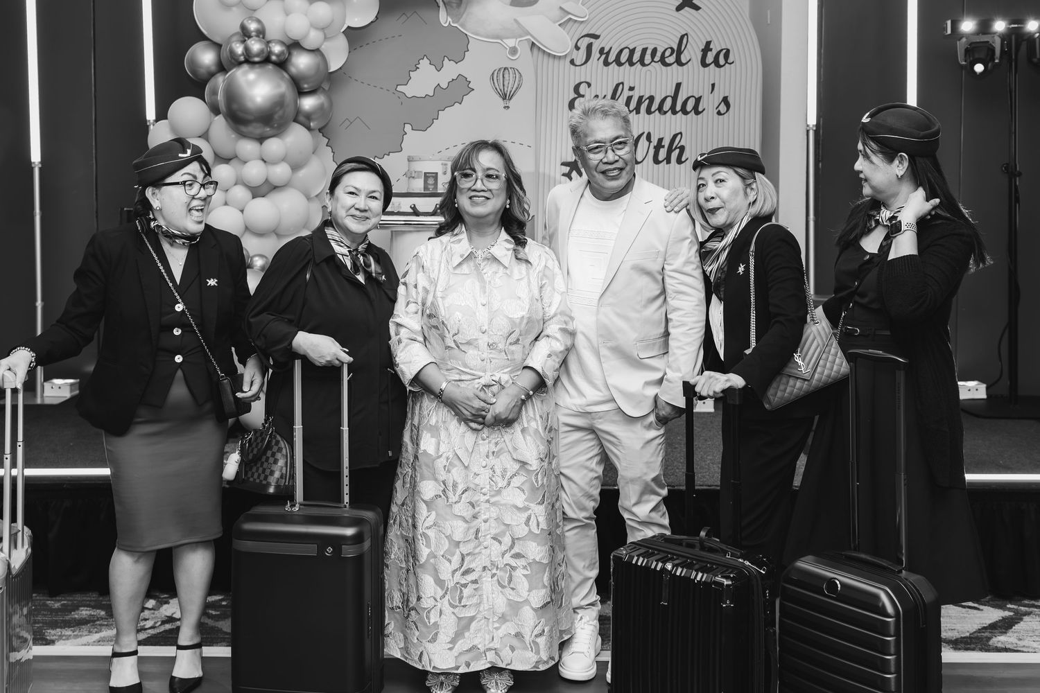 Group photo at Belinda's birthday party with travel theme, balloon backdrop, luggage props, and guests in flight attendant costumes.
