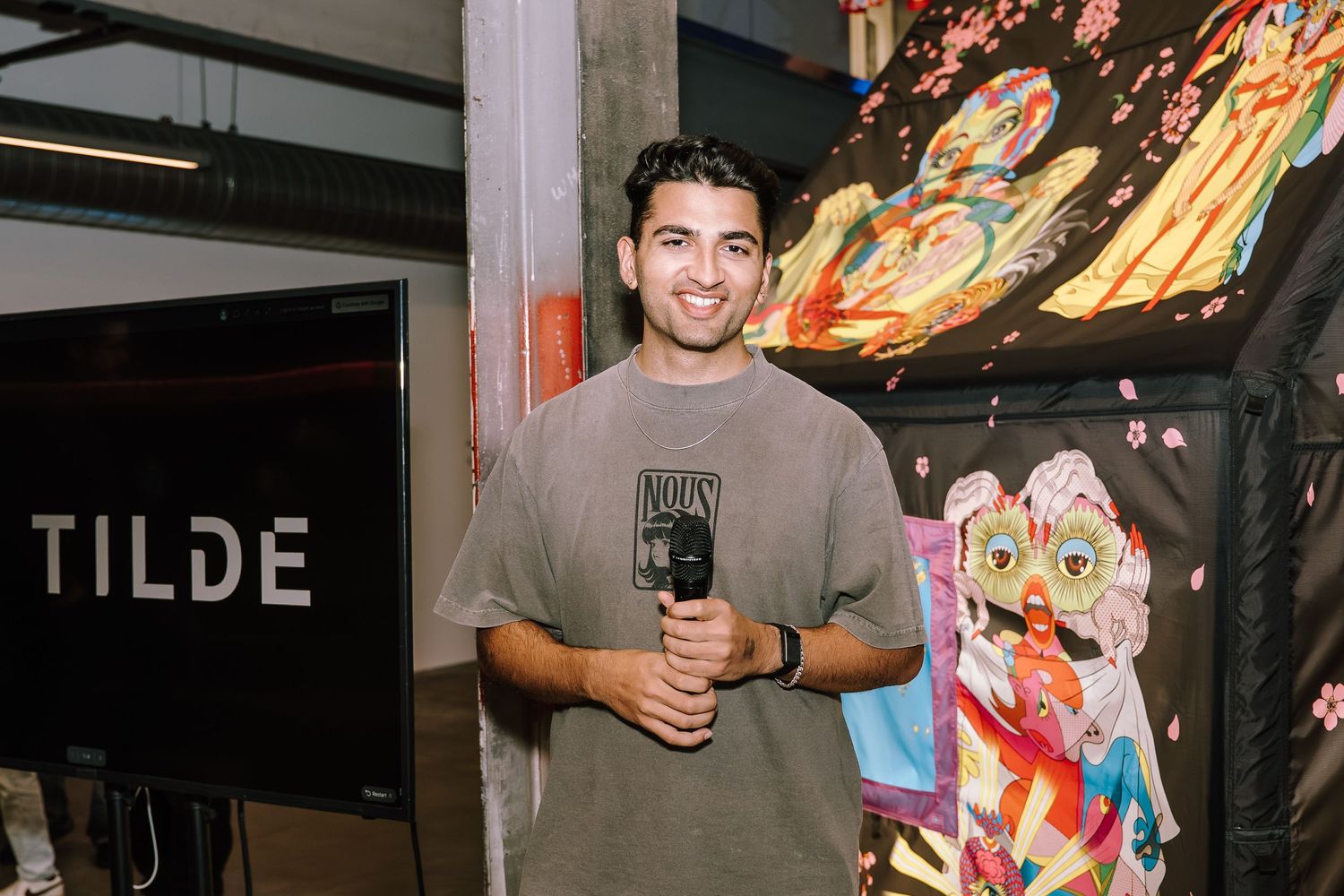 A young man smiling and holding a microphone at a TILDE event with colorful artwork in the background.