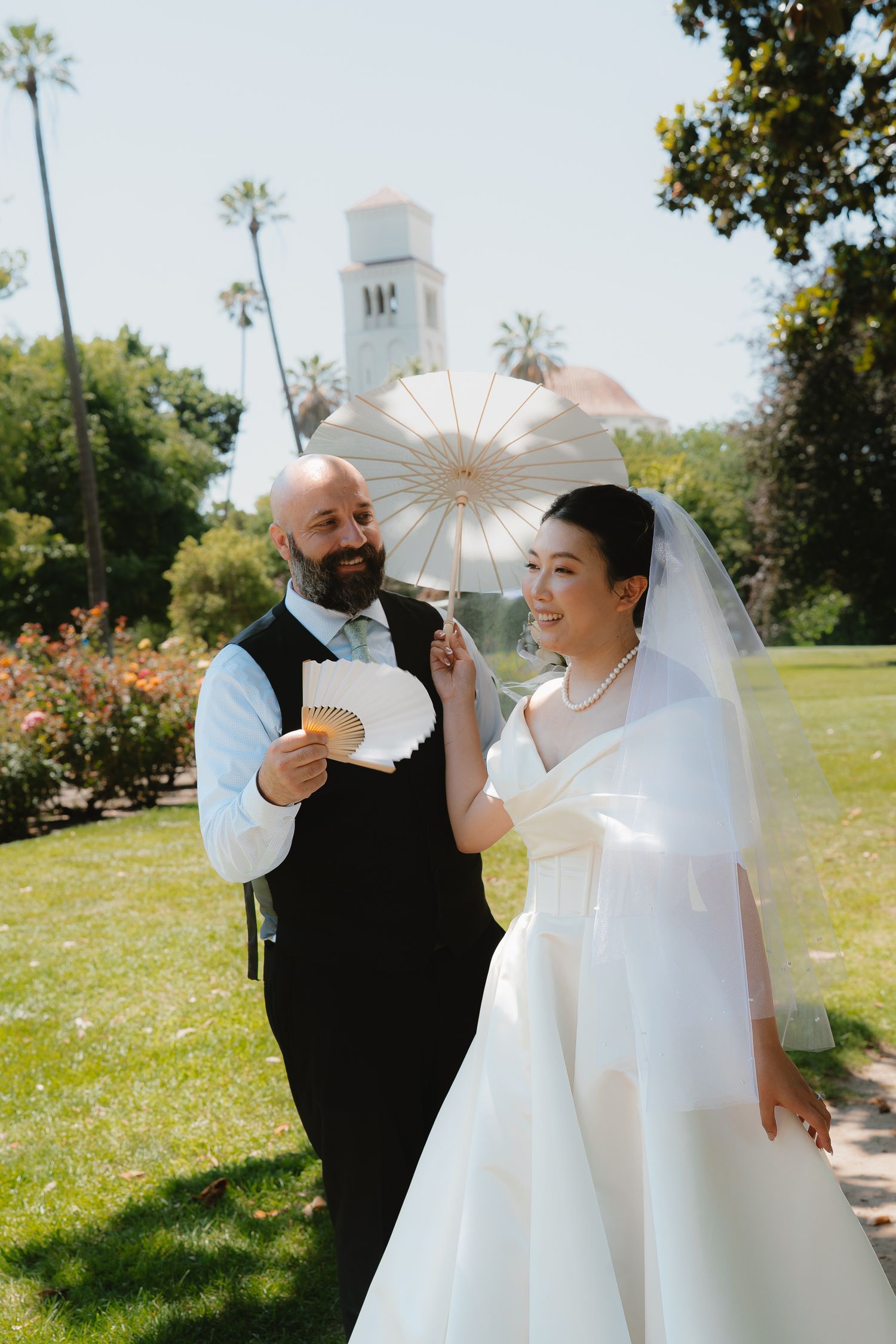 Bride and groom walk outdoors on sunny day, she holds white parasol while he fans himself with paper fan.