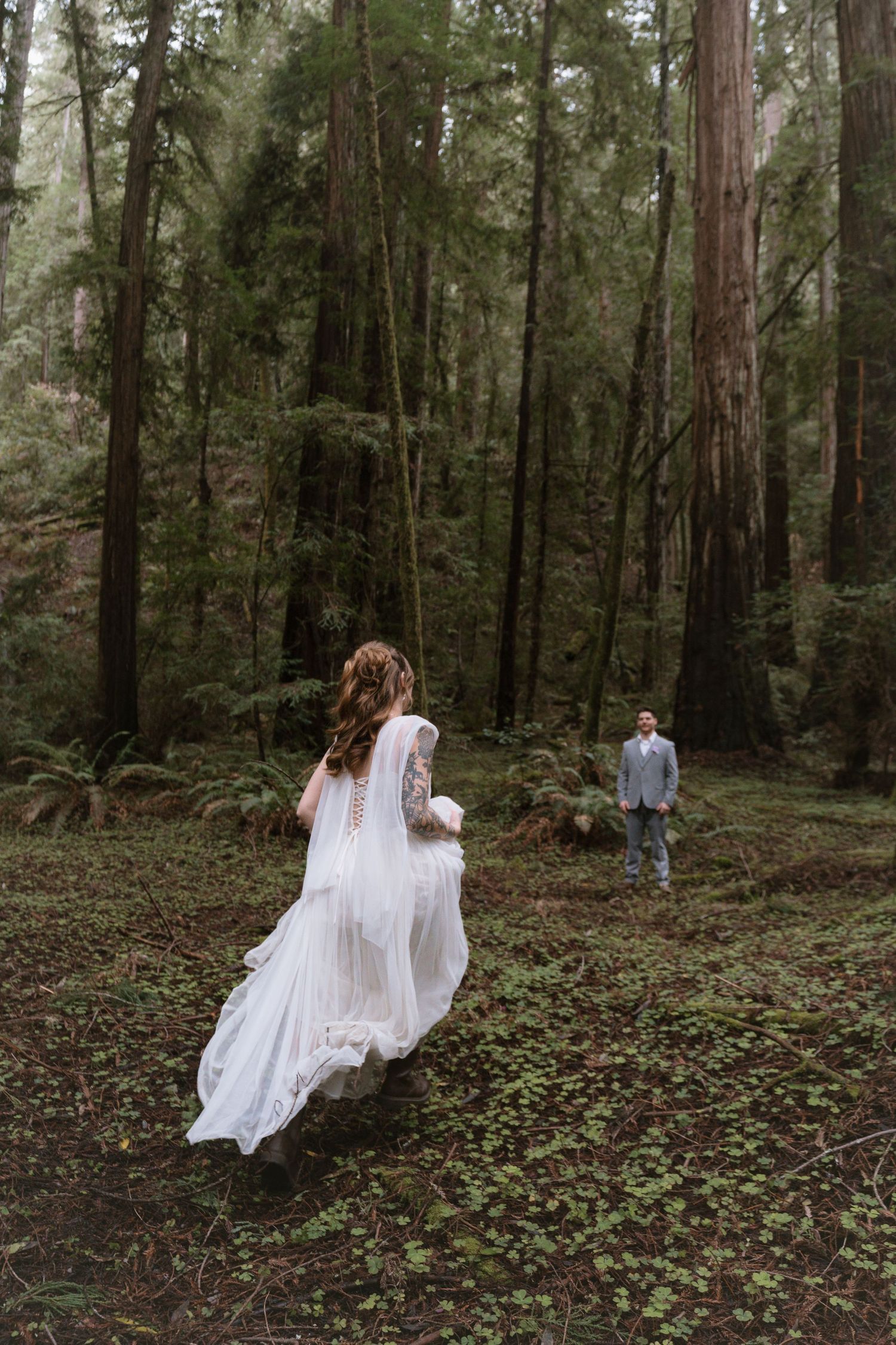 Bride in flowing white lace gown walks toward groom in misty redwood forest during romantic woodland elopement.