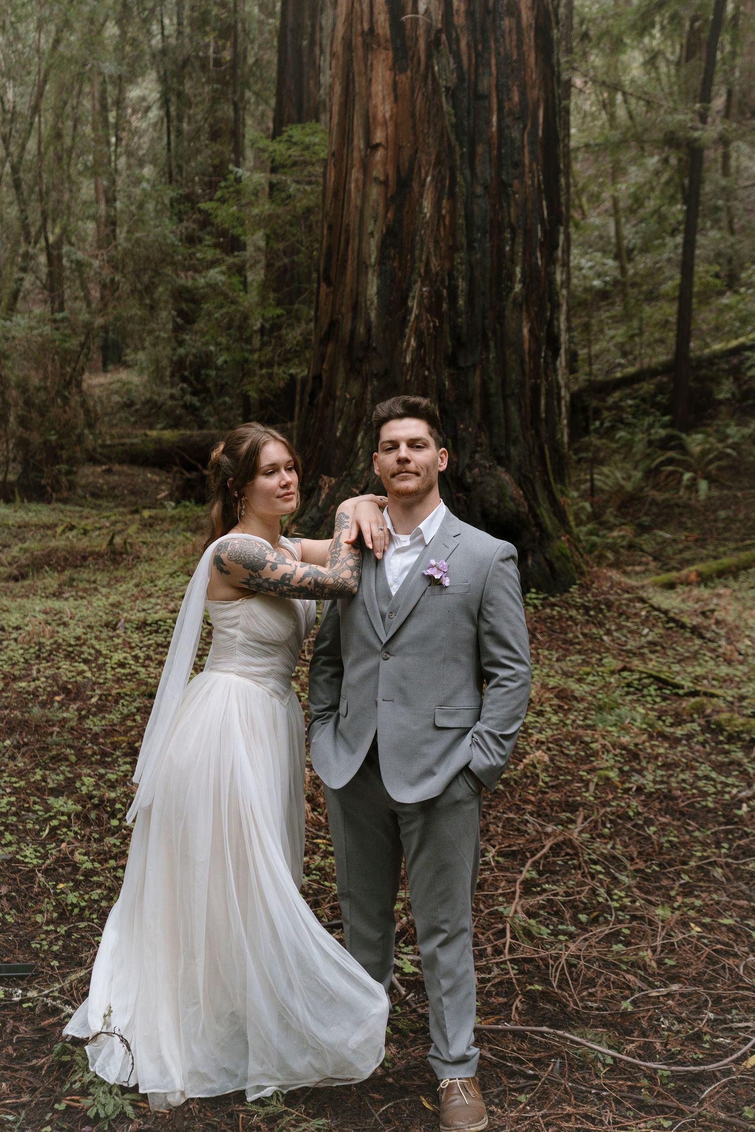 Bride and groom pose in a lush redwood forest, she in a flowing white gown, he in a grey suit.