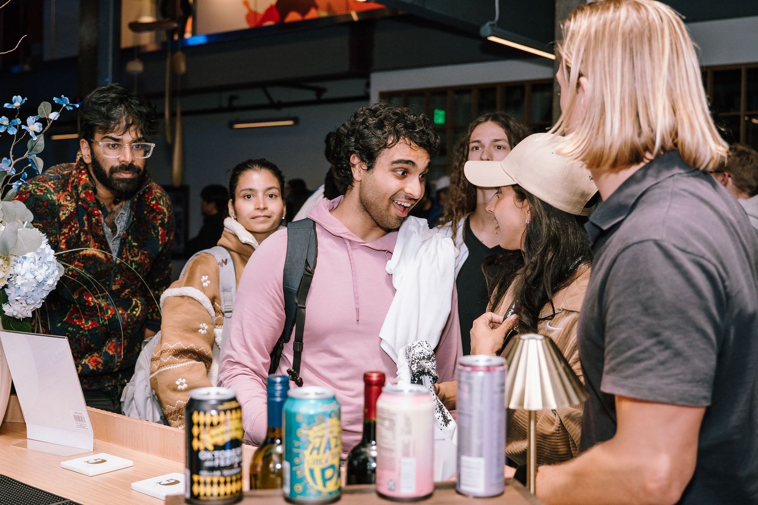 Group of young adults socializing at a bar counter with colorful canned drinks displayed on the counter.
