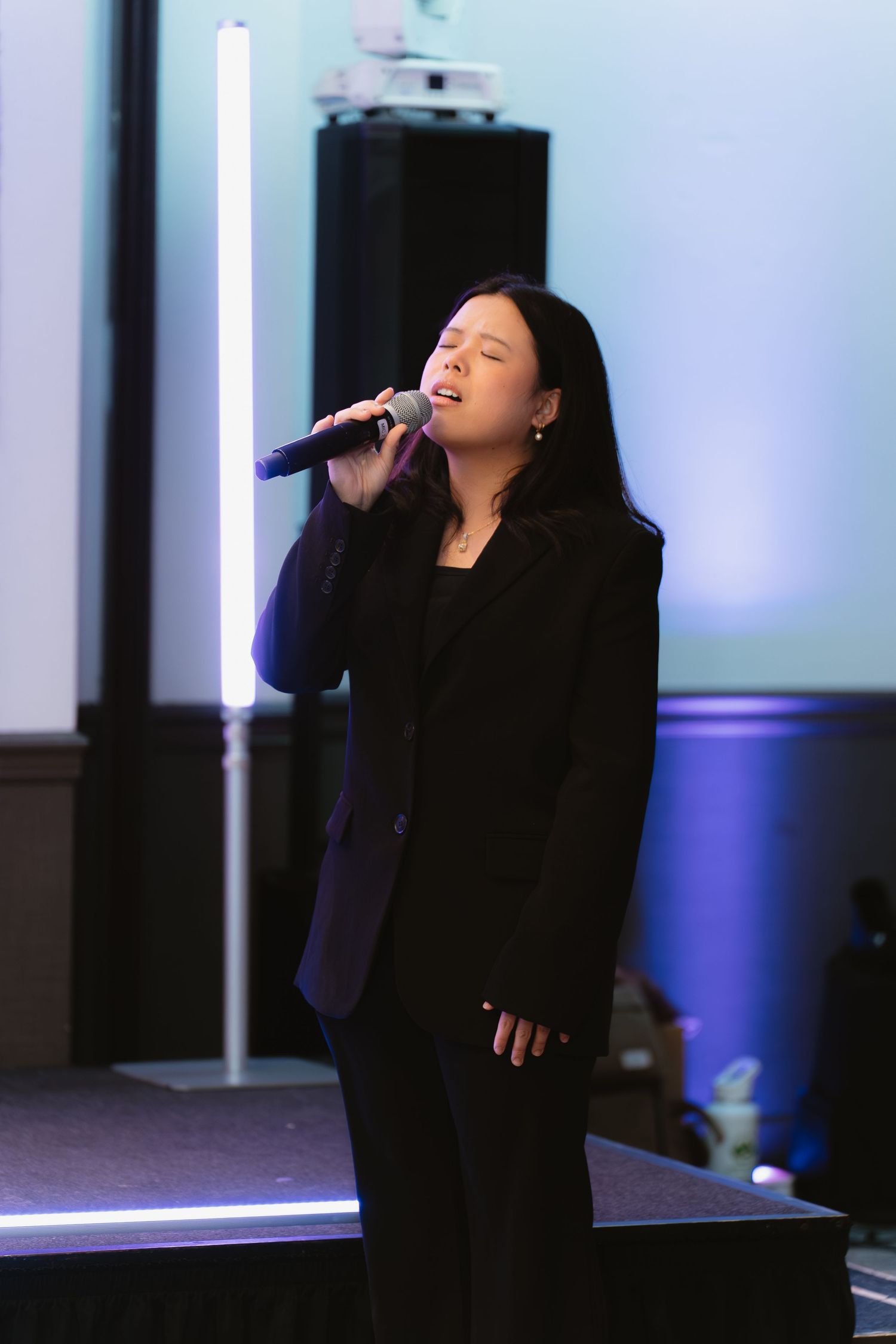 Young woman in black suit singing passionately into microphone on stage with blue lighting backdrop.