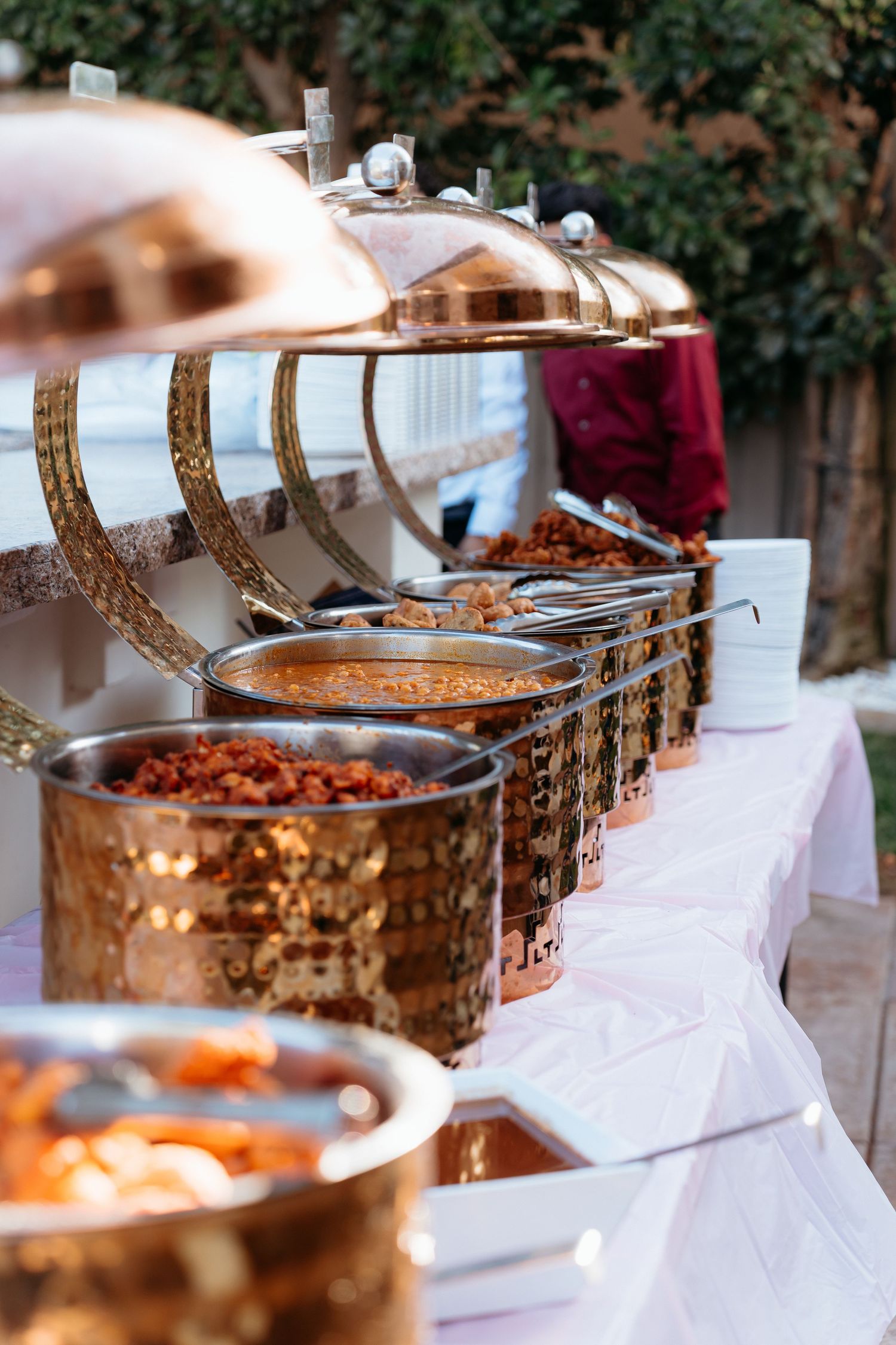 Elegant buffet setup with ornate copper chafing dishes filled with food on a white tablecloth at an outdoor event.