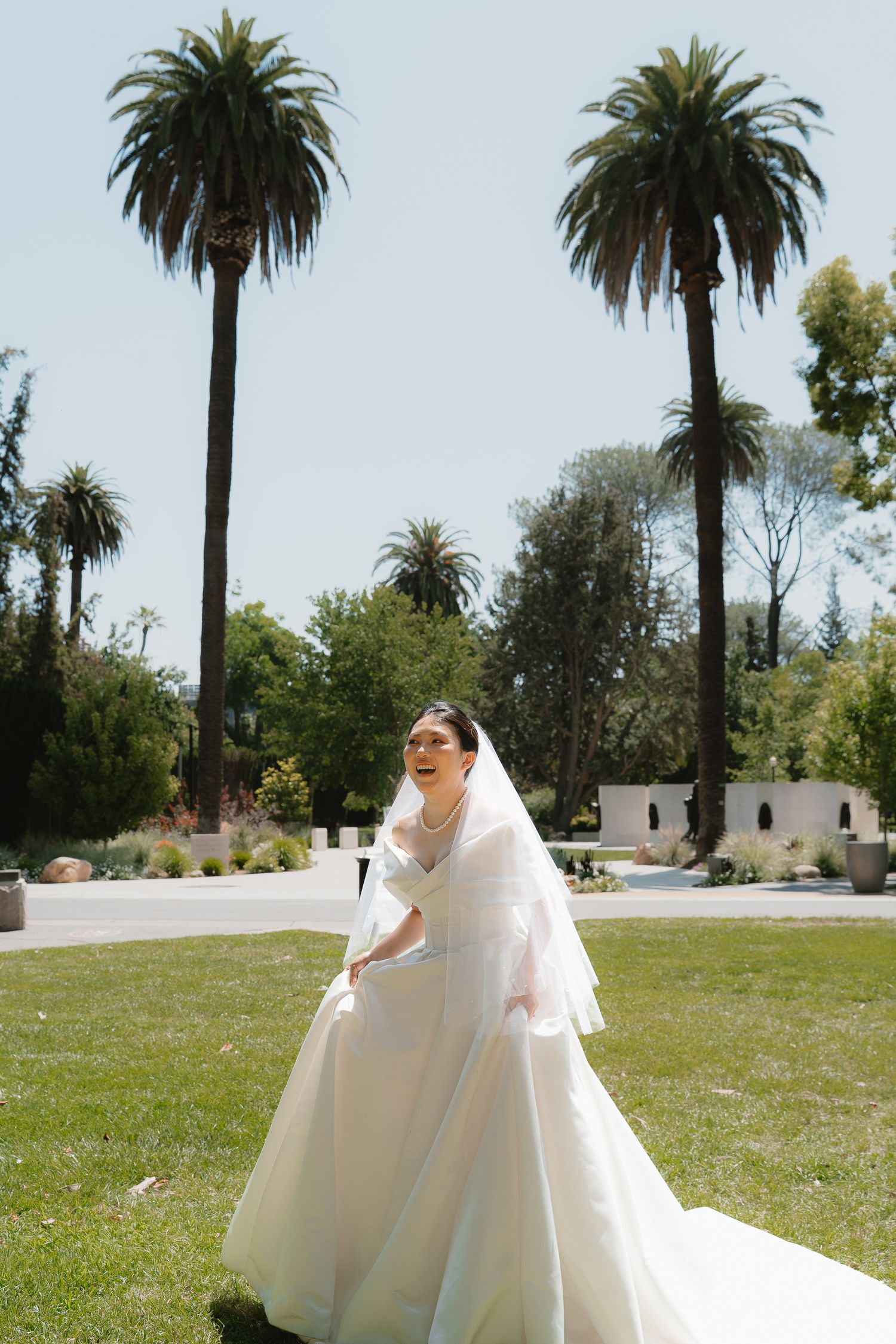 Bride in white ball gown and veil standing on green lawn with tall palm trees in sunny outdoor wedding setting.