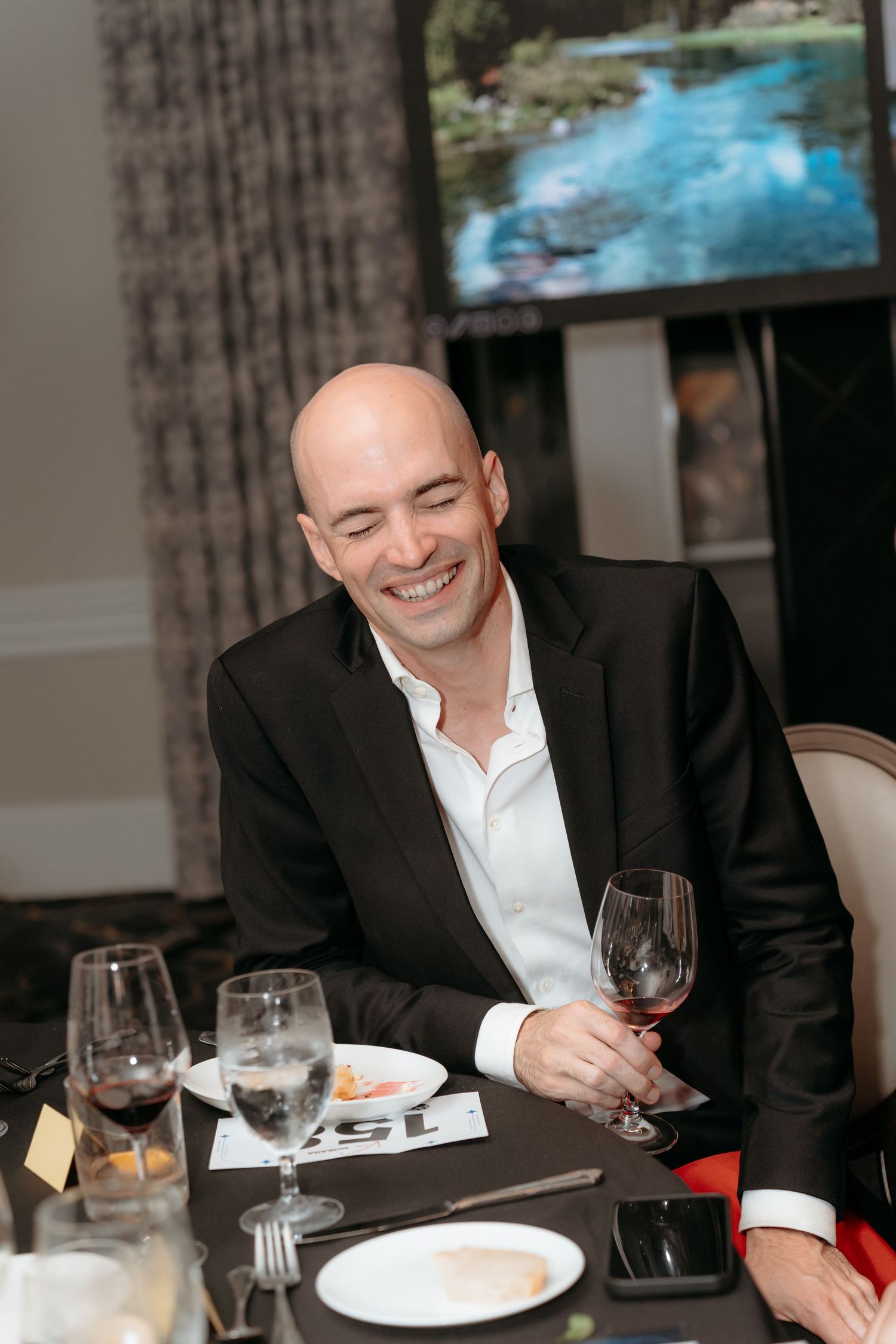 A person in a black suit jacket enjoying wine and dinner at an elegant restaurant table.