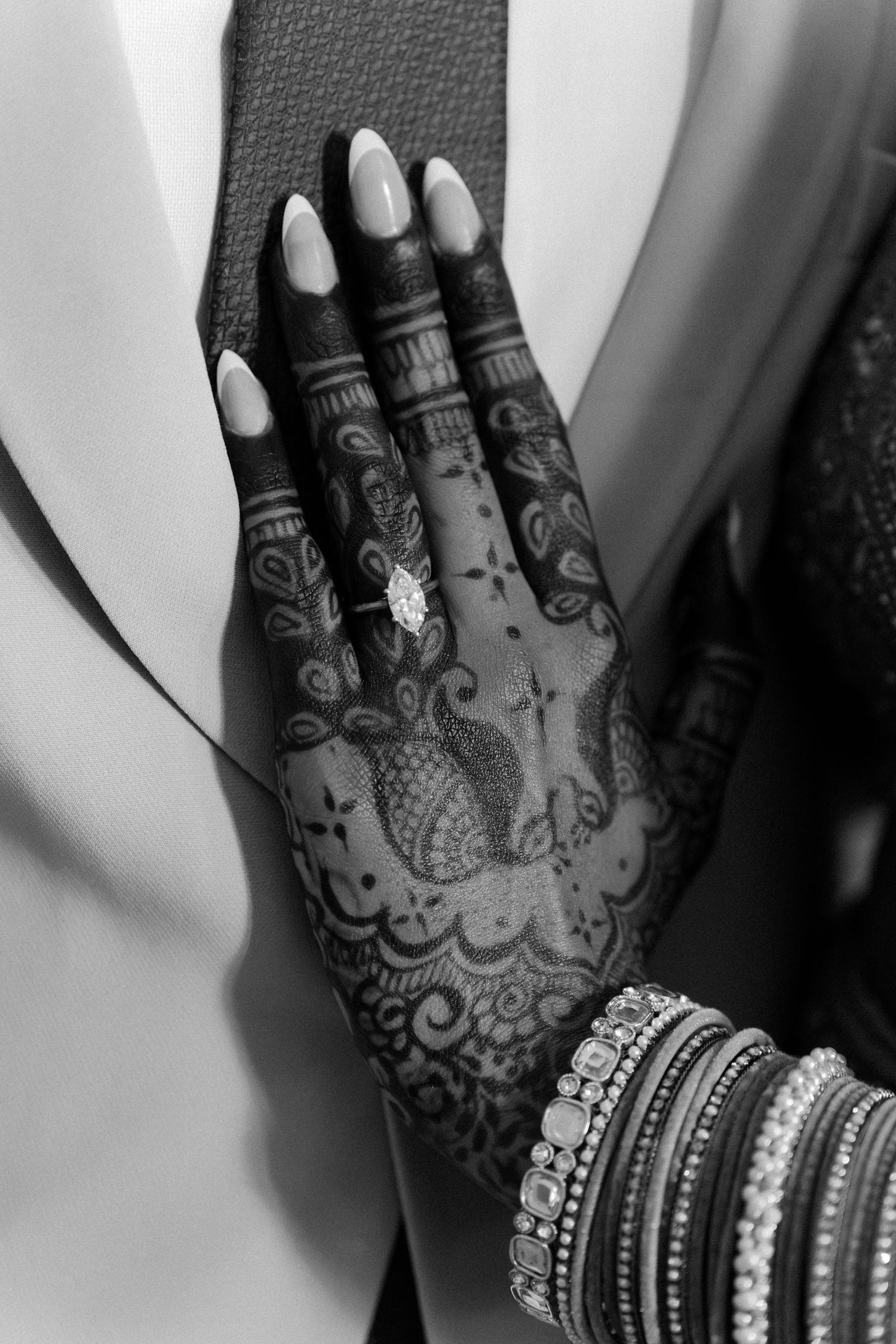 Black and white photo of a hand with intricate henna mehndi design, diamond ring, and bangles resting on a suit.