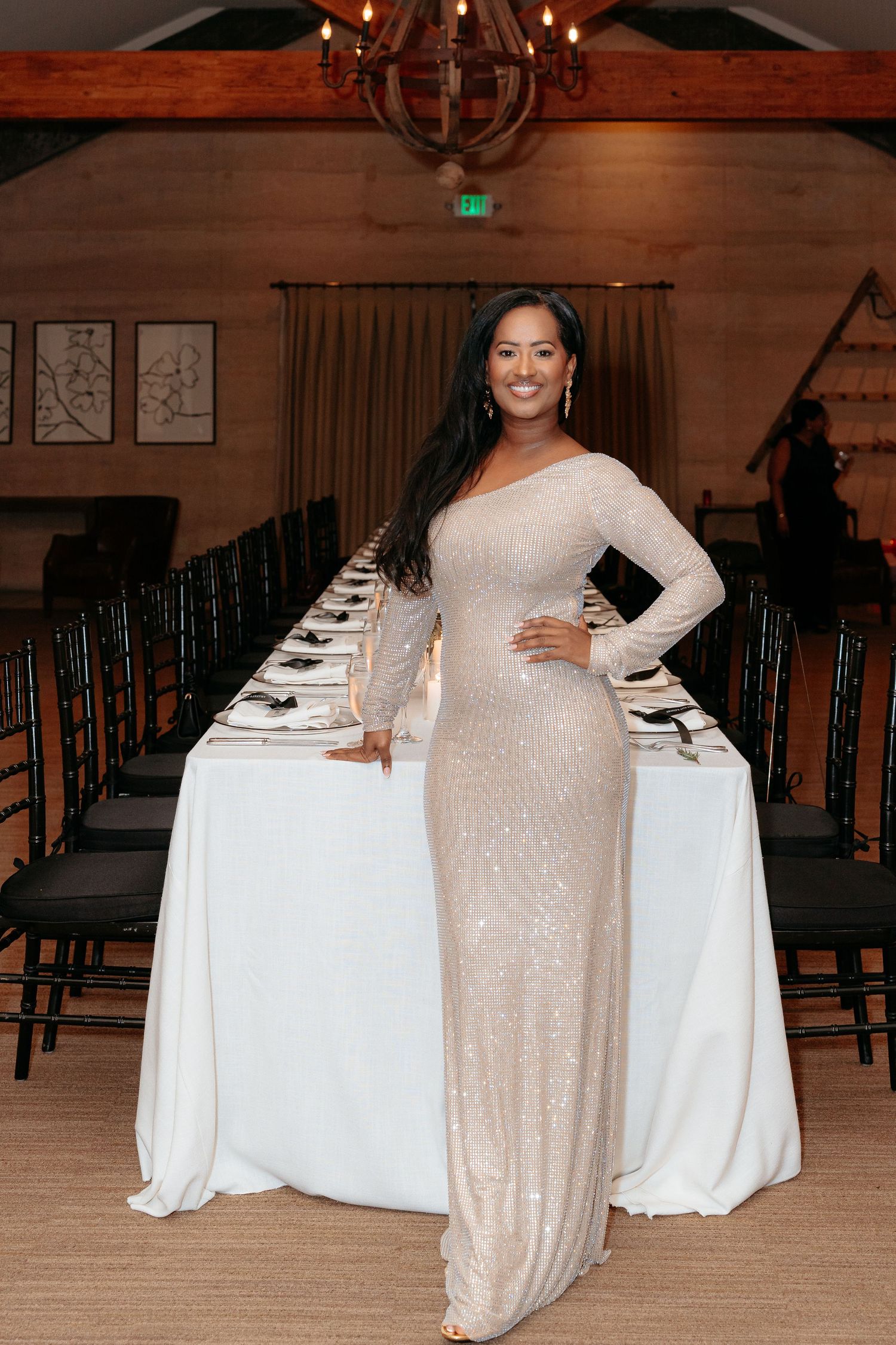 Woman in elegant sequin gown poses beside decorated banquet table in upscale event venue.