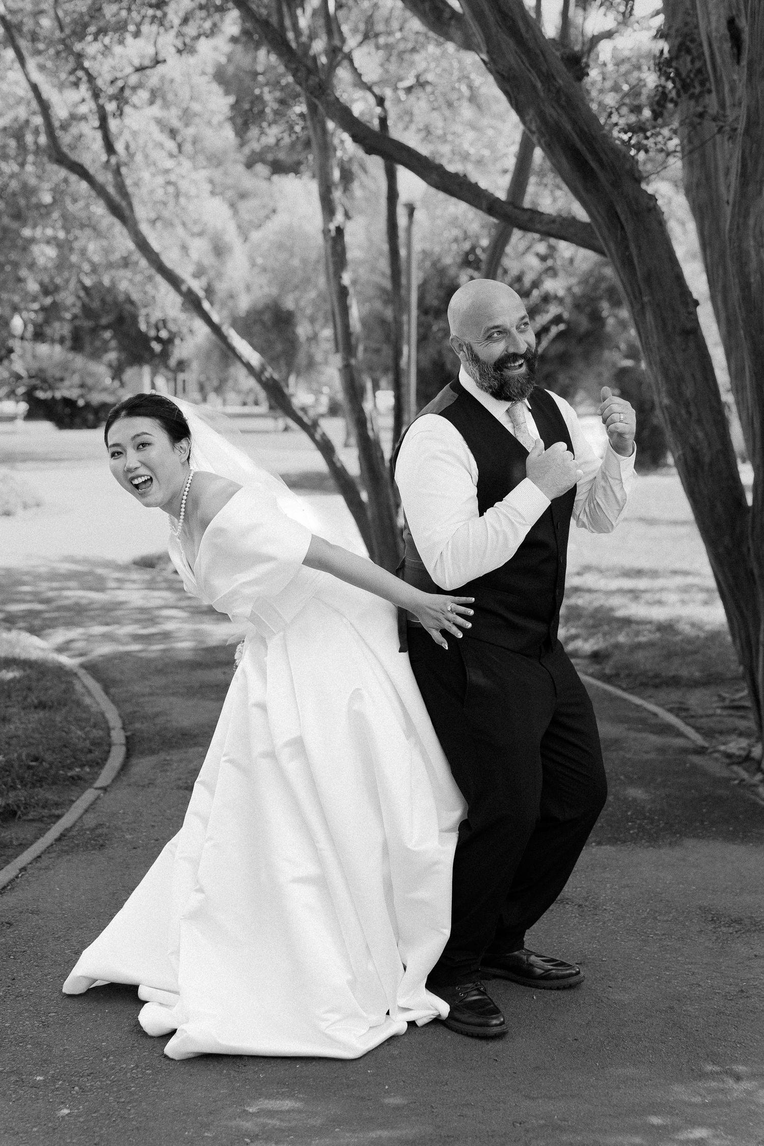 Joyful bride and groom dancing outdoors in black and white wedding photo among trees.