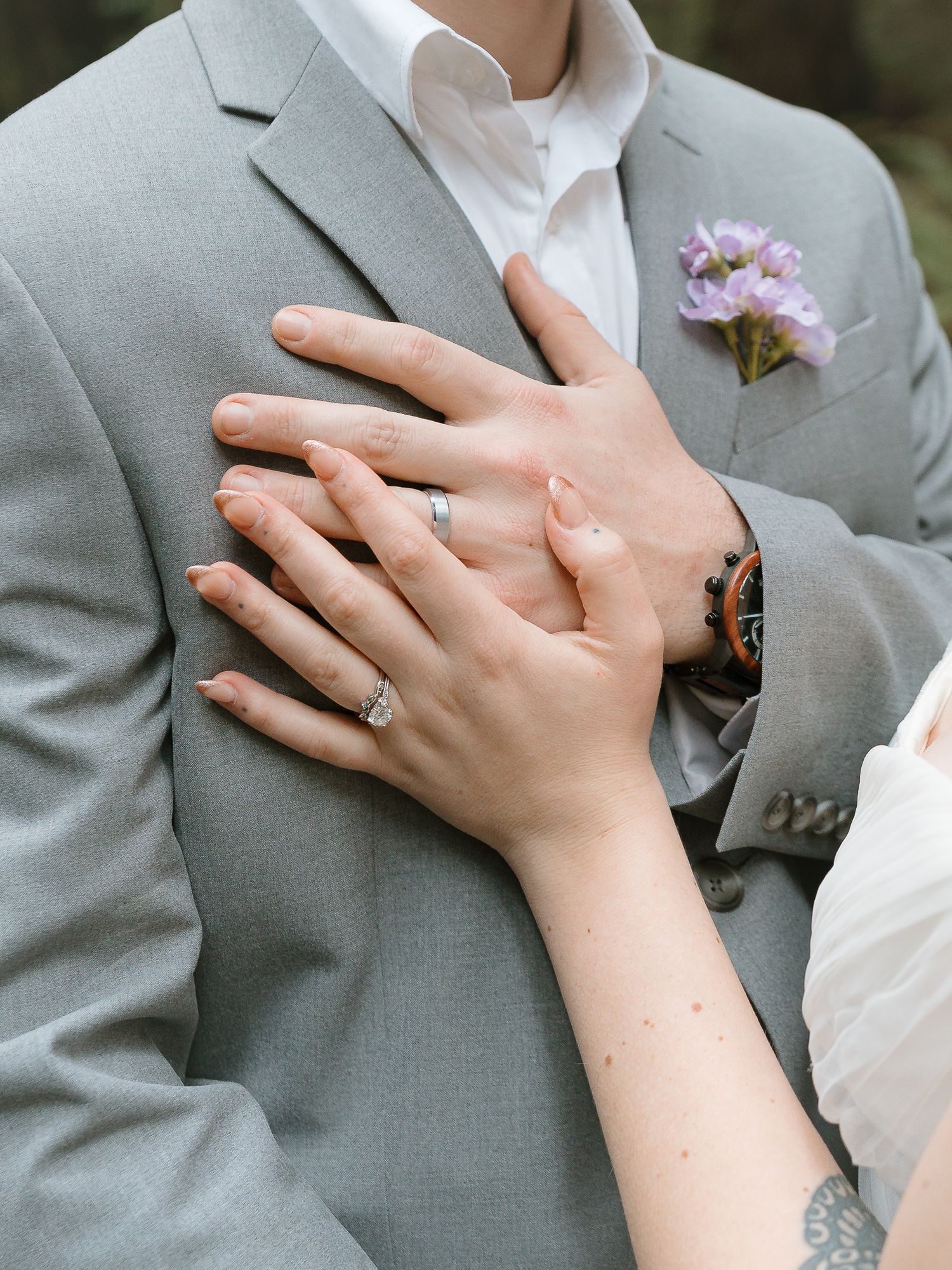 Bride and groom holding hands showing wedding rings, groom in gray suit with purple floral boutonniere.