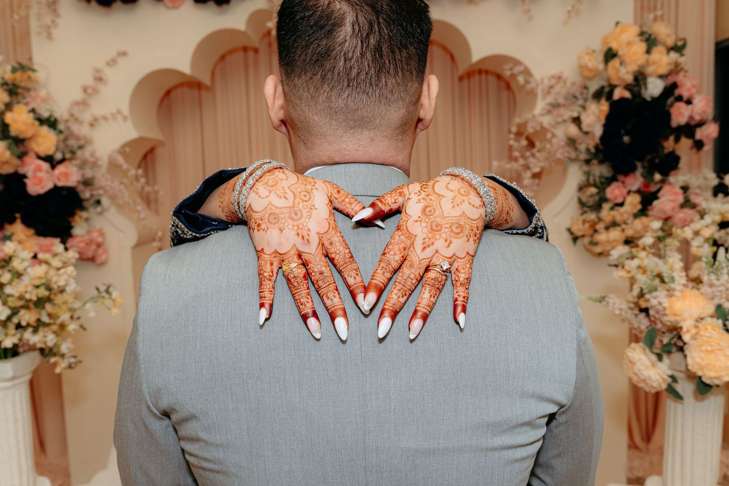 Bride's henna-adorned hands resting on groom's gray suit jacket at a wedding ceremony.