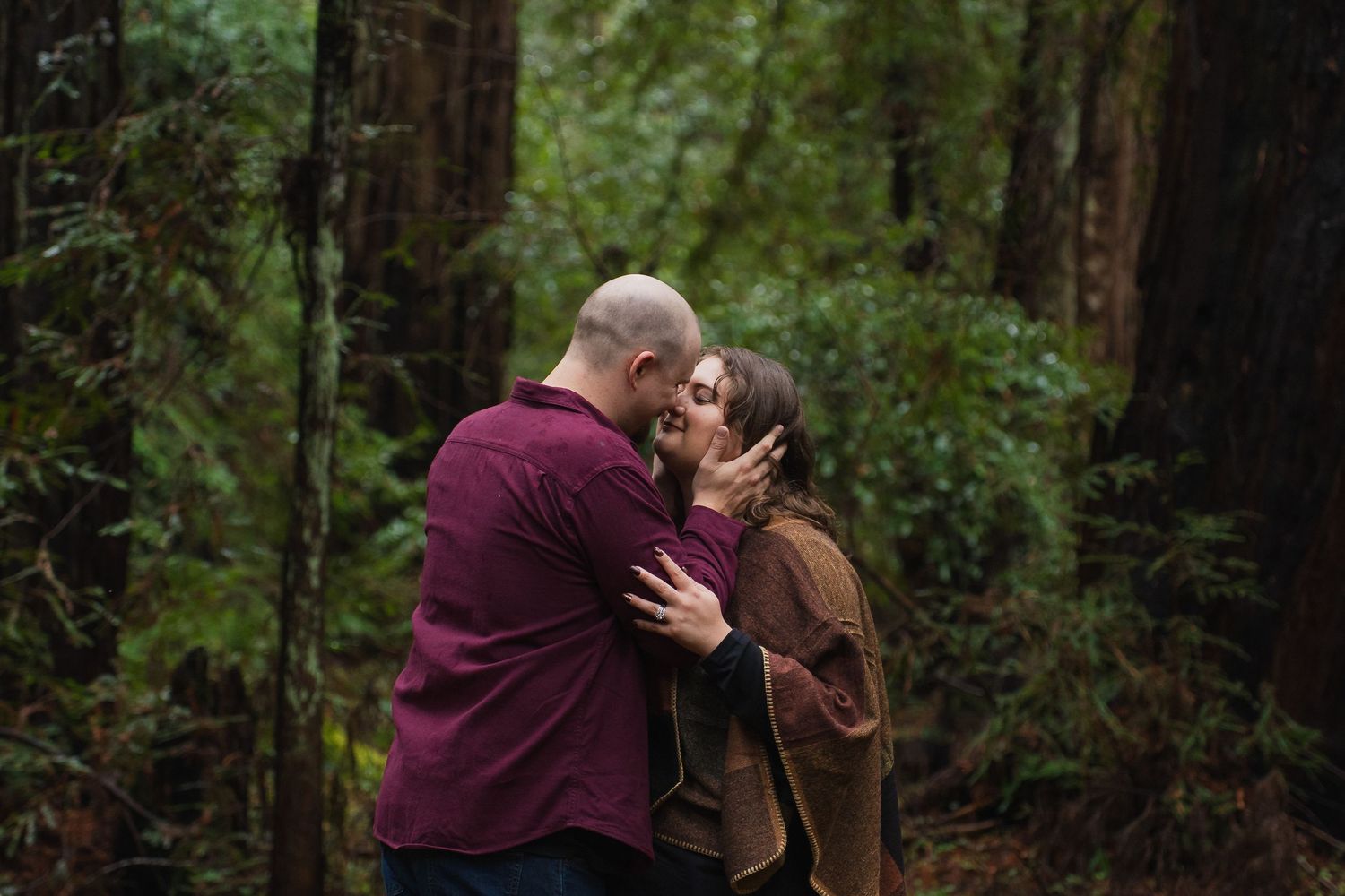 Couple sharing a romantic kiss surrounded by tall redwood trees in a lush green forest setting.