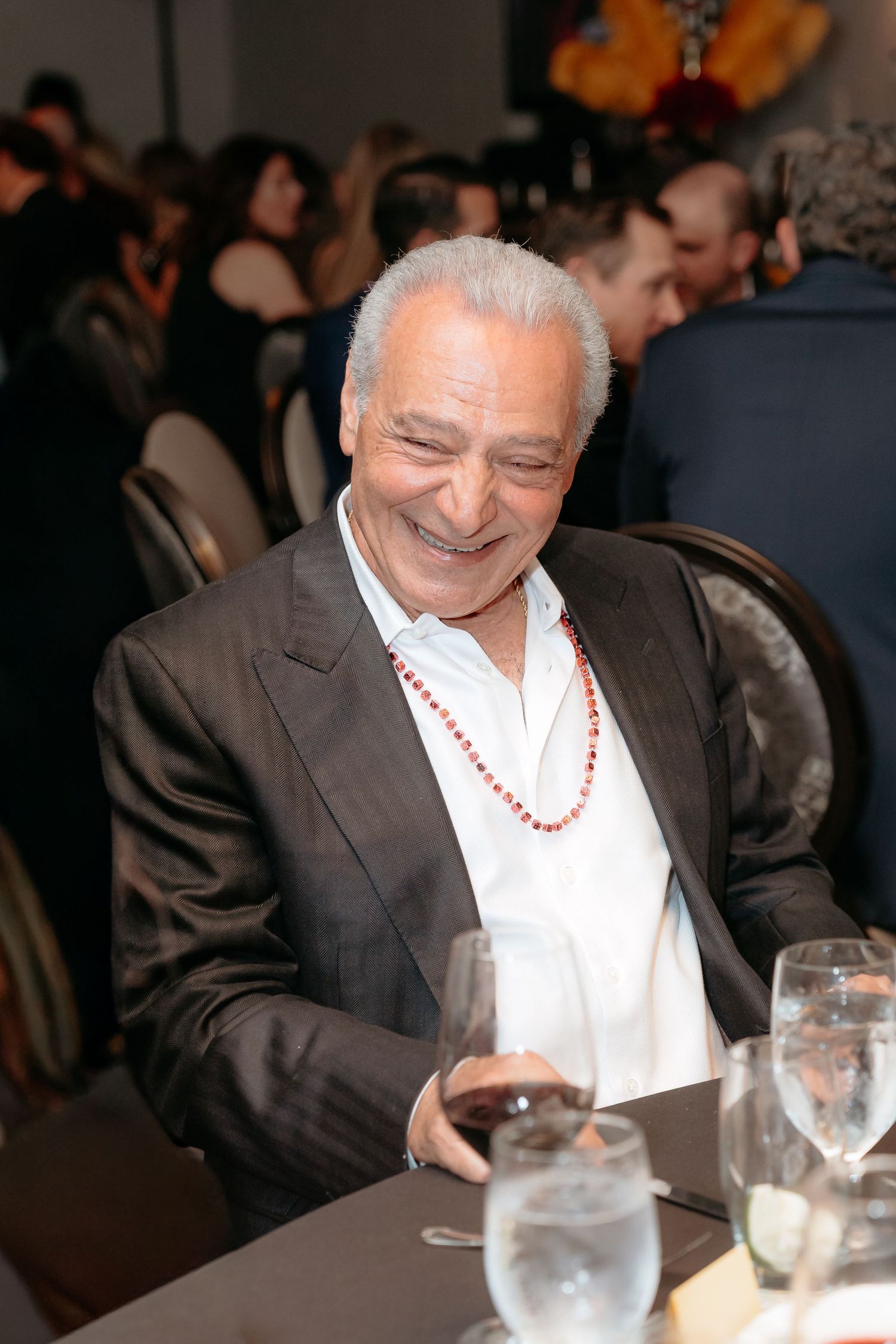 A person in a dark suit and white shirt smiles while sitting at a formal dining table with glassware.
