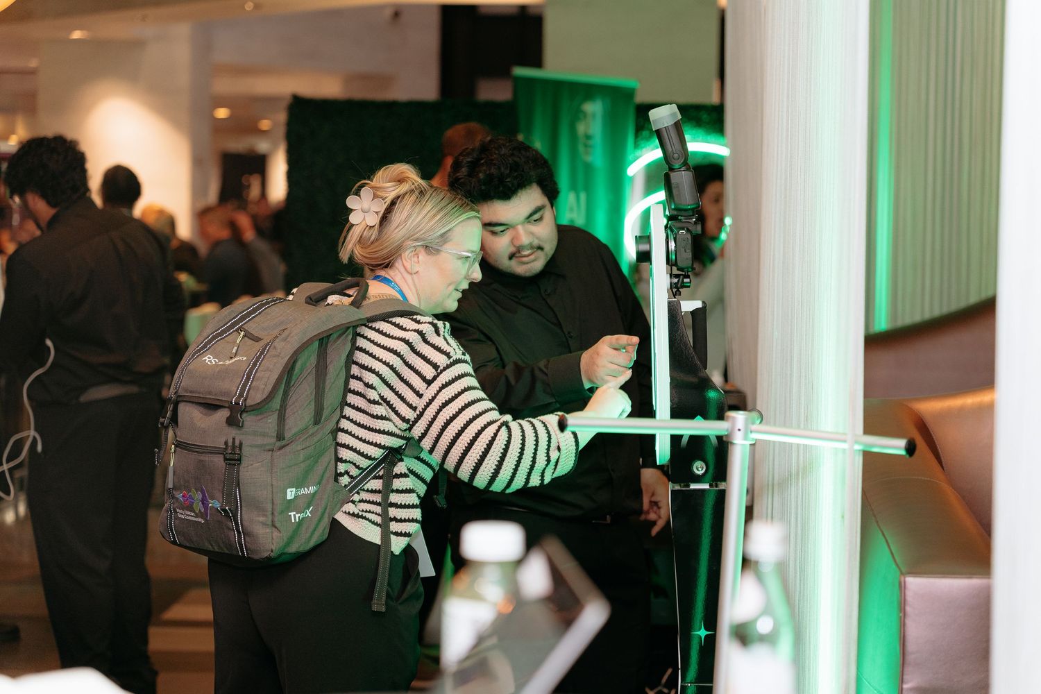Two people interact with a digital kiosk at a green-lit event venue with attendees in the background.