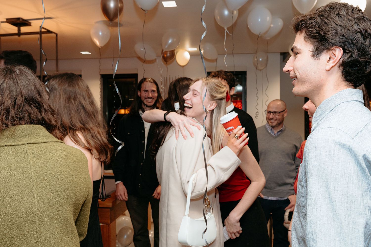 People celebrating at a party with white balloons, laughing and socializing indoors in a warmly lit venue.