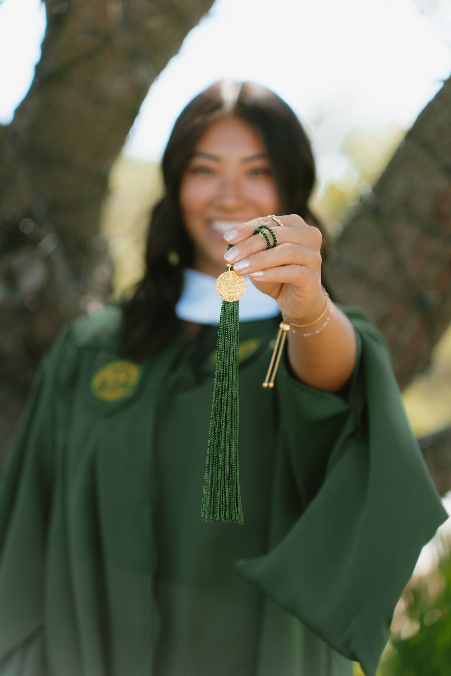 Smiling graduate in green cap and gown holding tassel medallion toward camera outdoors.