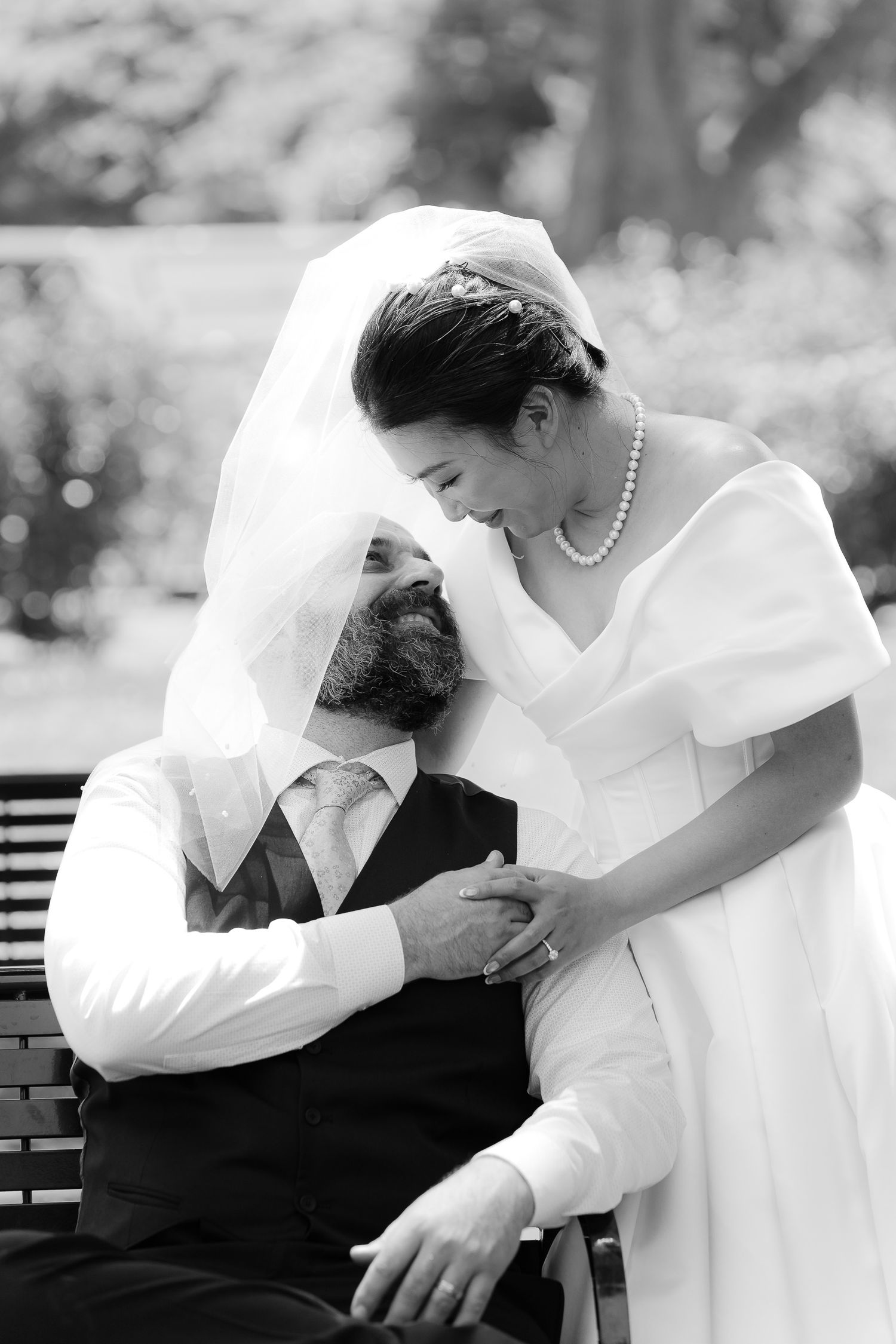 Black and white photo of bride and groom sharing tender moment on bench, bride's veil draped over groom.