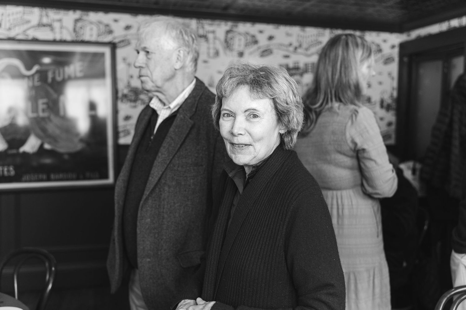 Black and white photo of an elderly couple at an indoor gathering with a poster on the stone wall behind them.