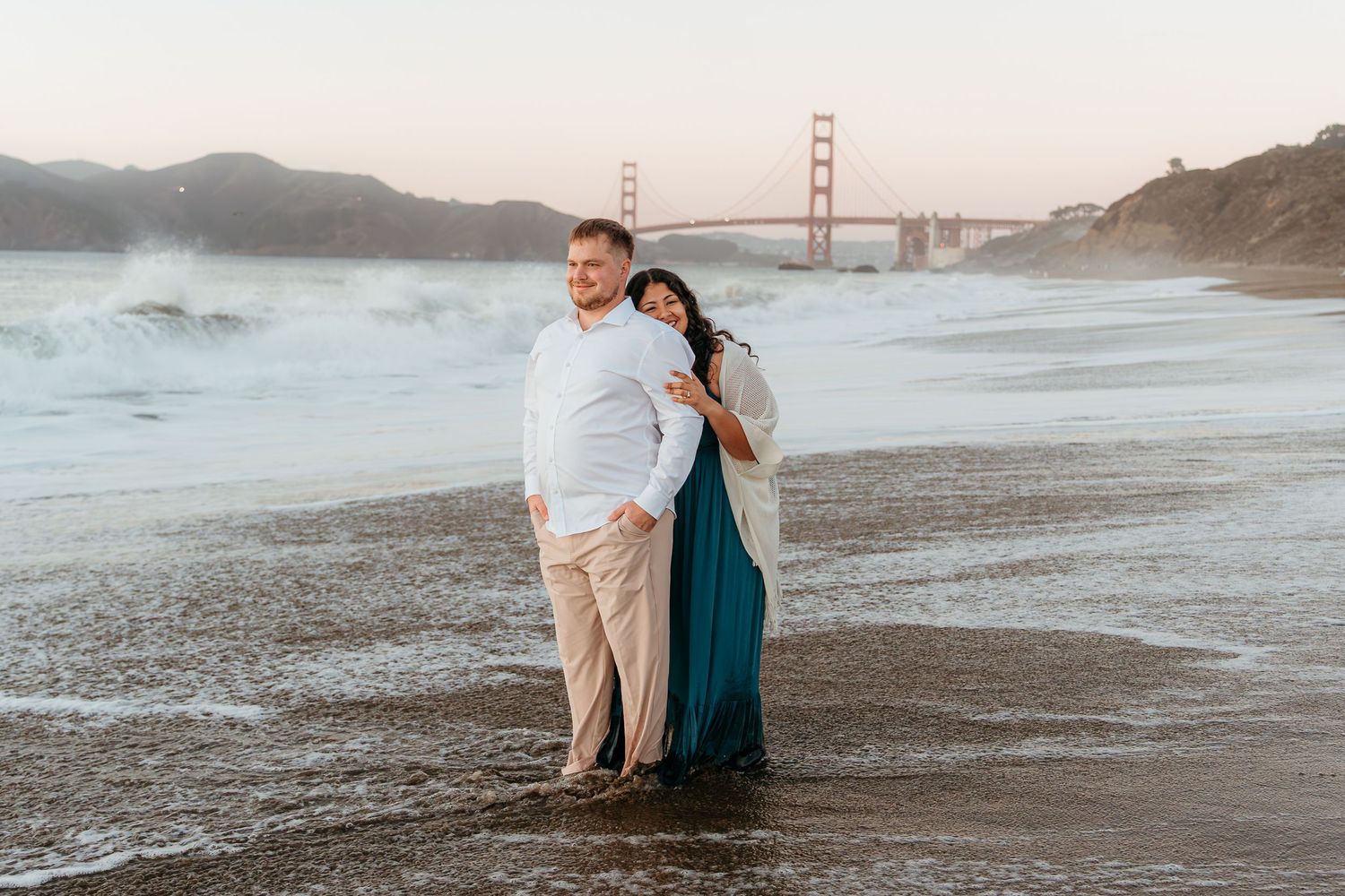 Couple embracing on Baker Beach with Golden Gate Bridge backdrop at sunset in San Francisco.