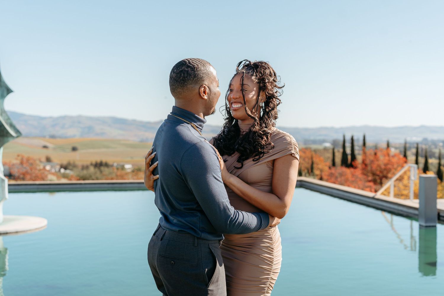 Couple embracing and smiling by an infinity pool with scenic mountain and vineyard views on a clear sunny day.