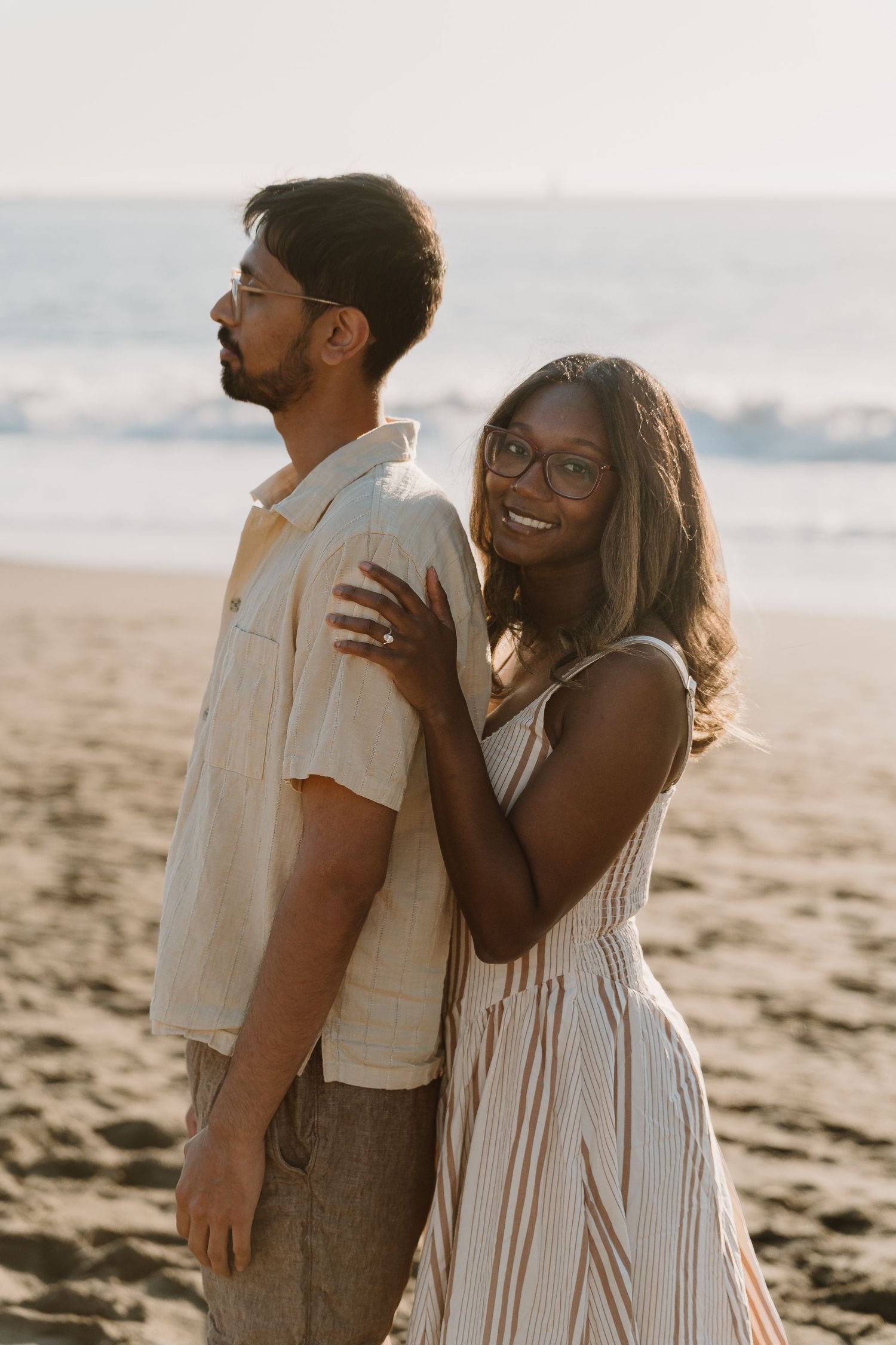 Couple posing on a sandy beach at sunset, woman in striped dress smiling at camera, man in linen shirt looking away.