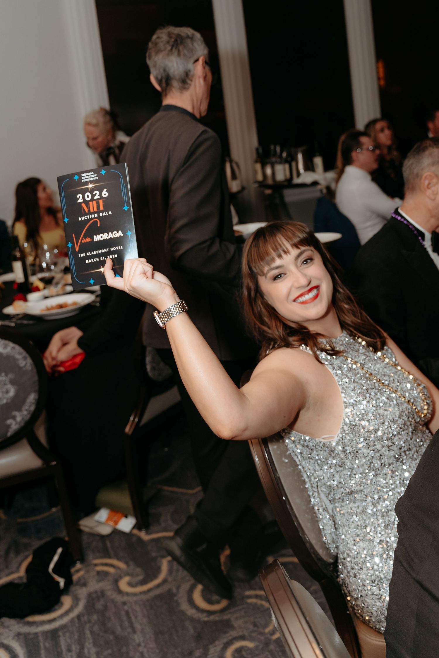 Woman in silver sequin dress holding a 2026 AHF Auction Gala award at an elegant event dinner.