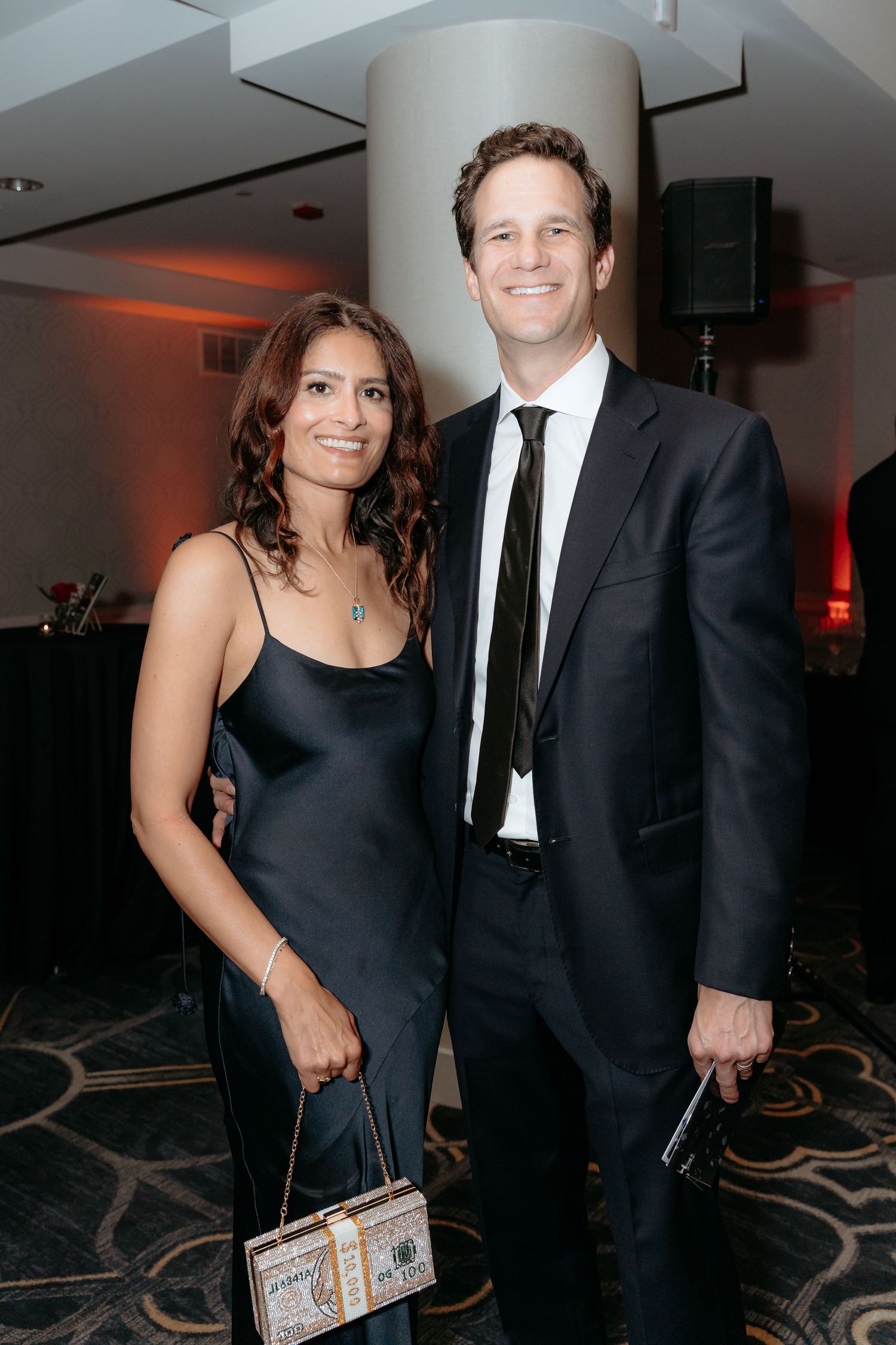A couple in formal attire poses at an evening event, with the woman wearing a black dress and holding a clutch.