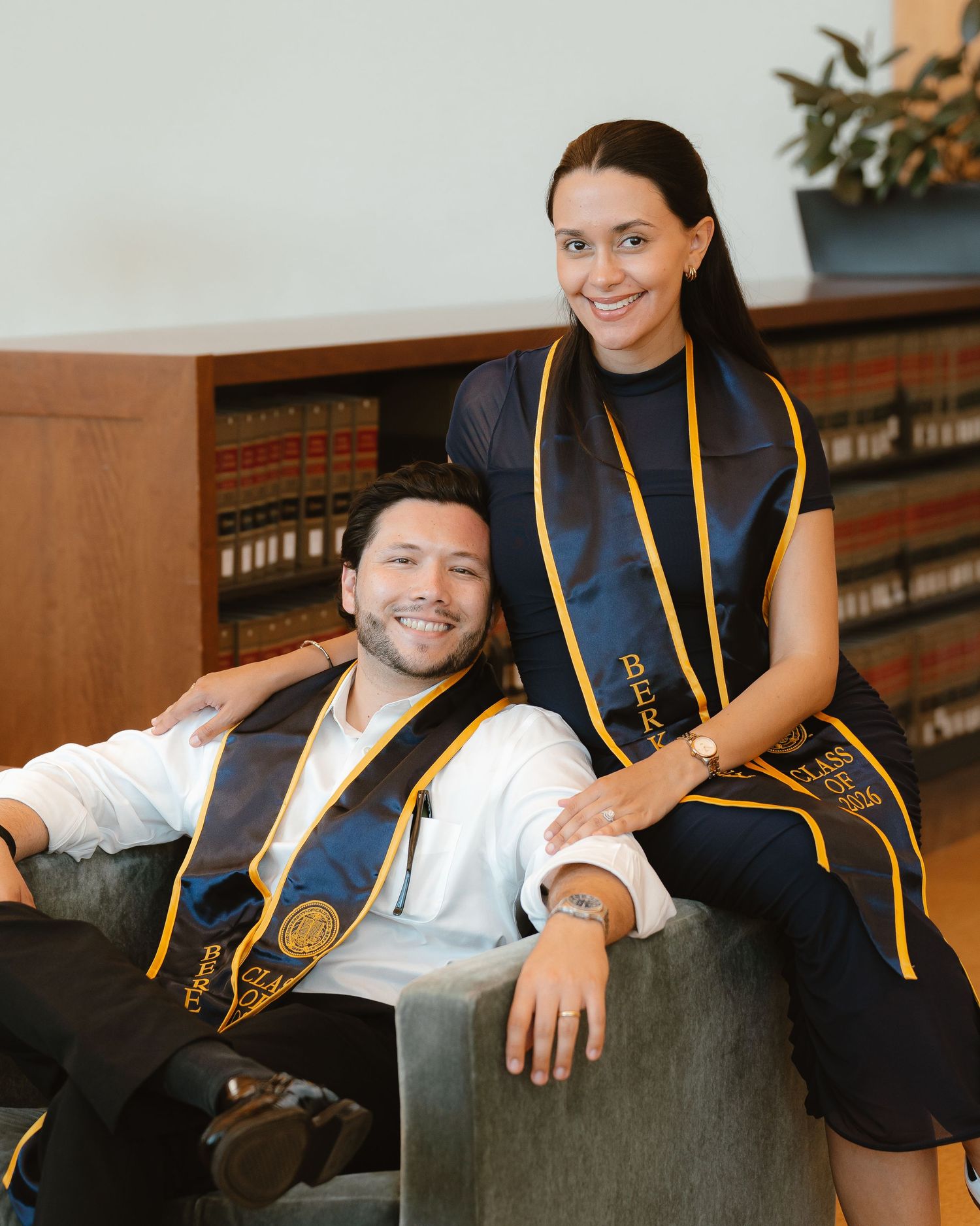 Two graduates wearing navy and gold stoles smile proudly for a photo in front of a bookshelf.