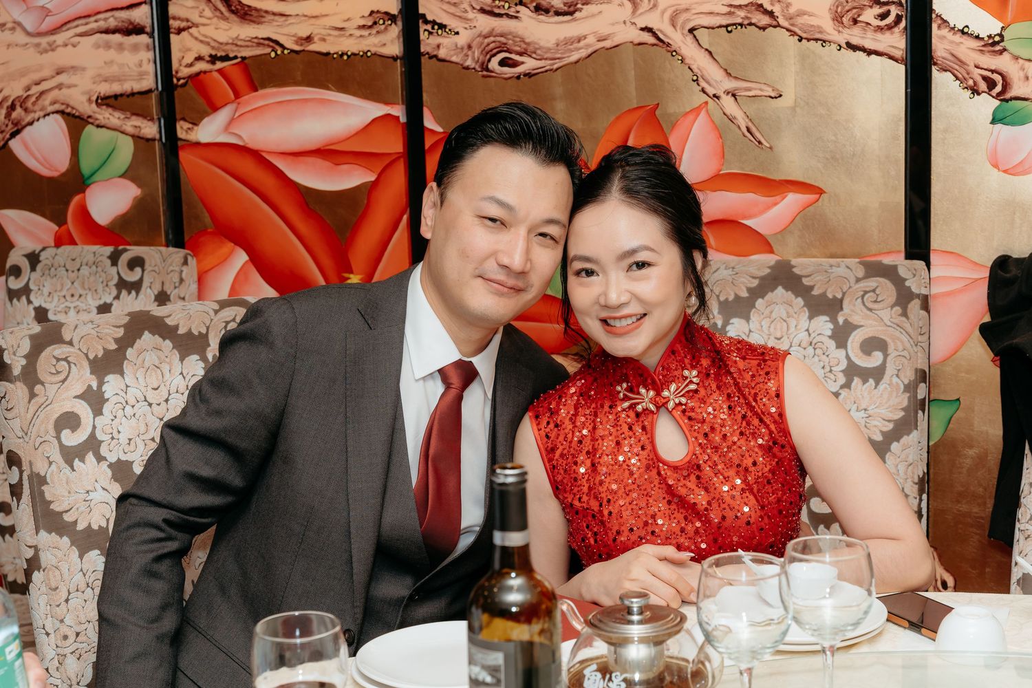 Couple smiling together at a Chinese wedding banquet, woman in red qipao and man in gray suit.