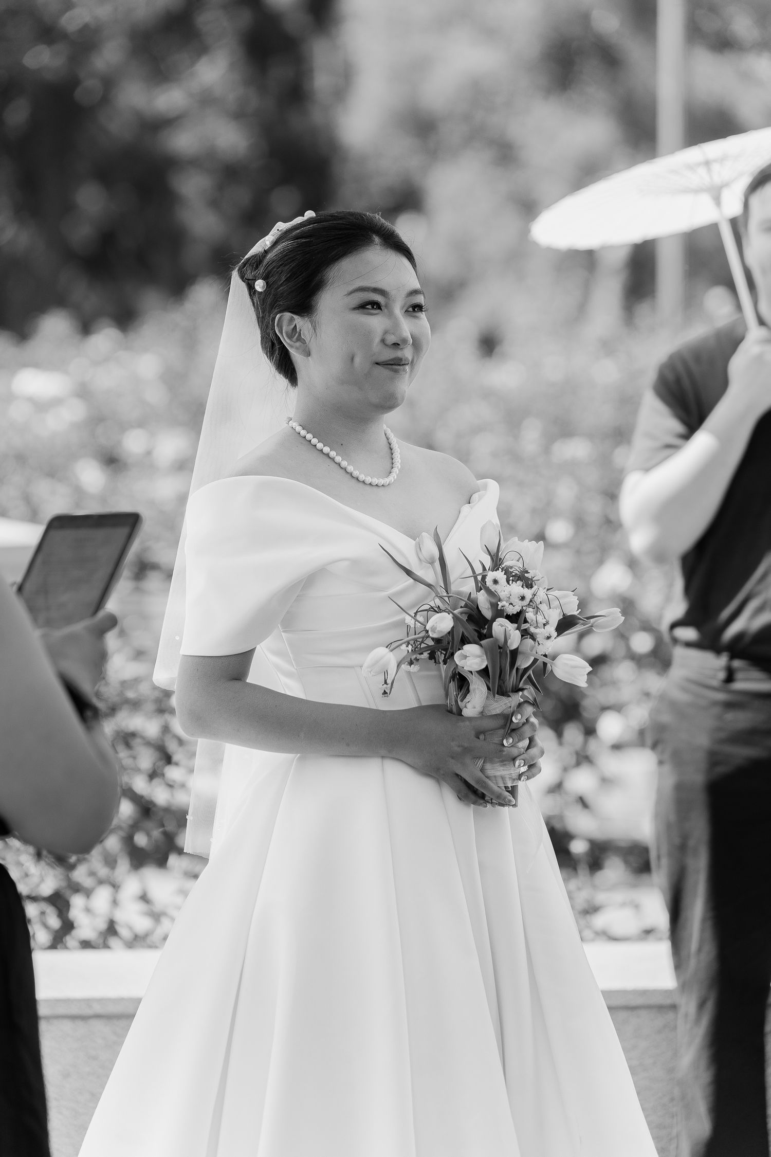 Black and white photo of a smiling Asian bride in a white gown holding a bouquet at an outdoor wedding ceremony.