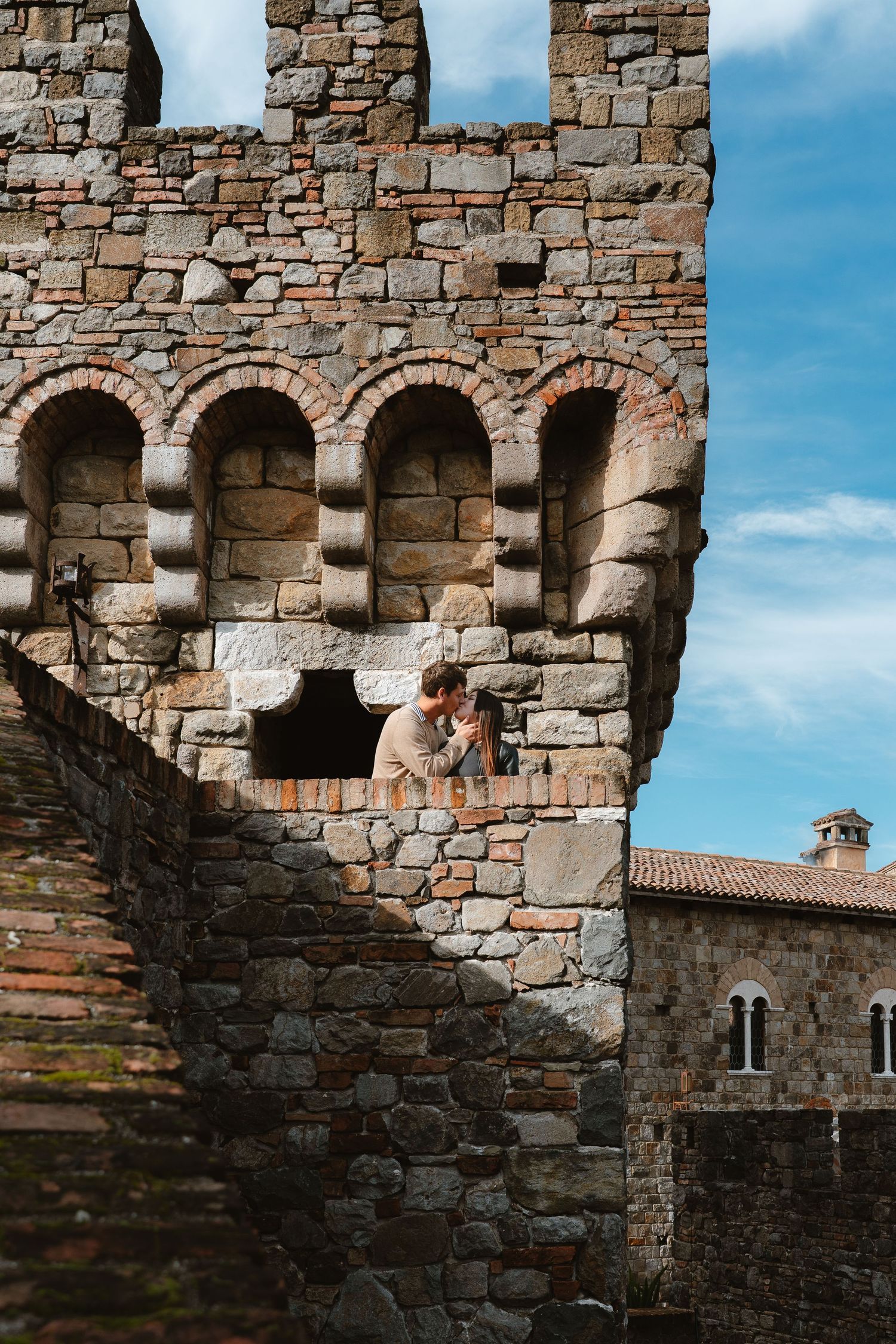A couple shares an intimate moment against the backdrop of ancient stone castle walls on a sunny day.