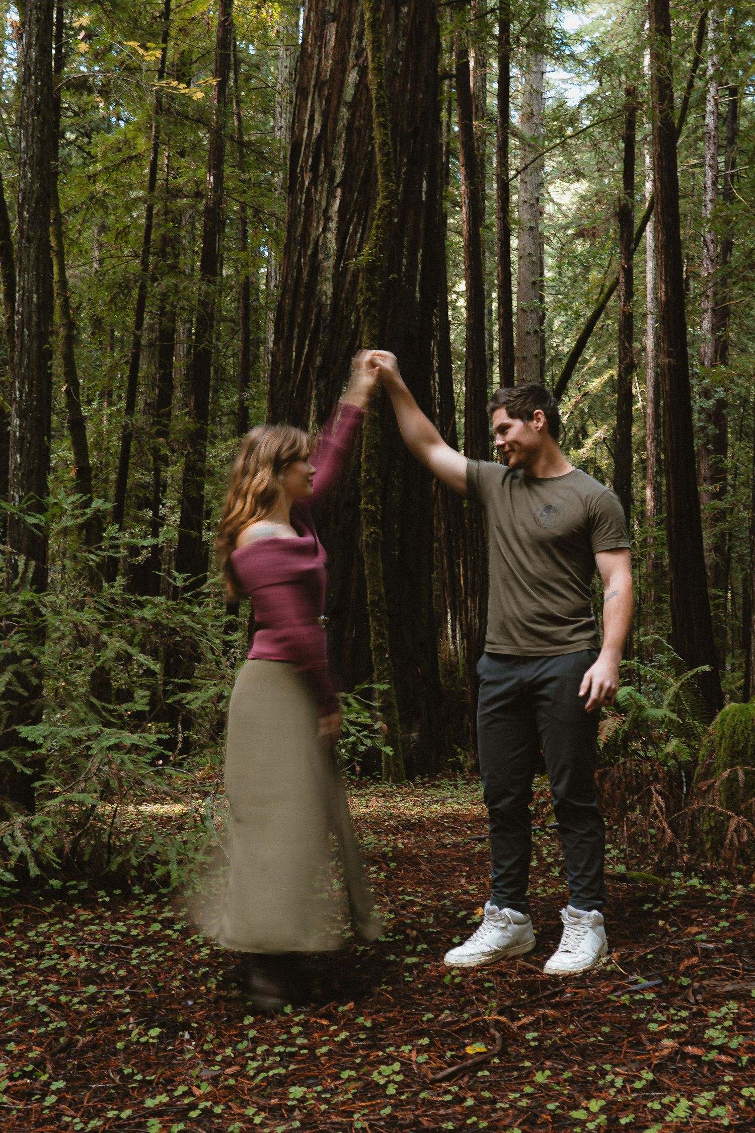 A couple dances together in a lush redwood forest, the man spinning the woman in a maxi skirt among towering trees.