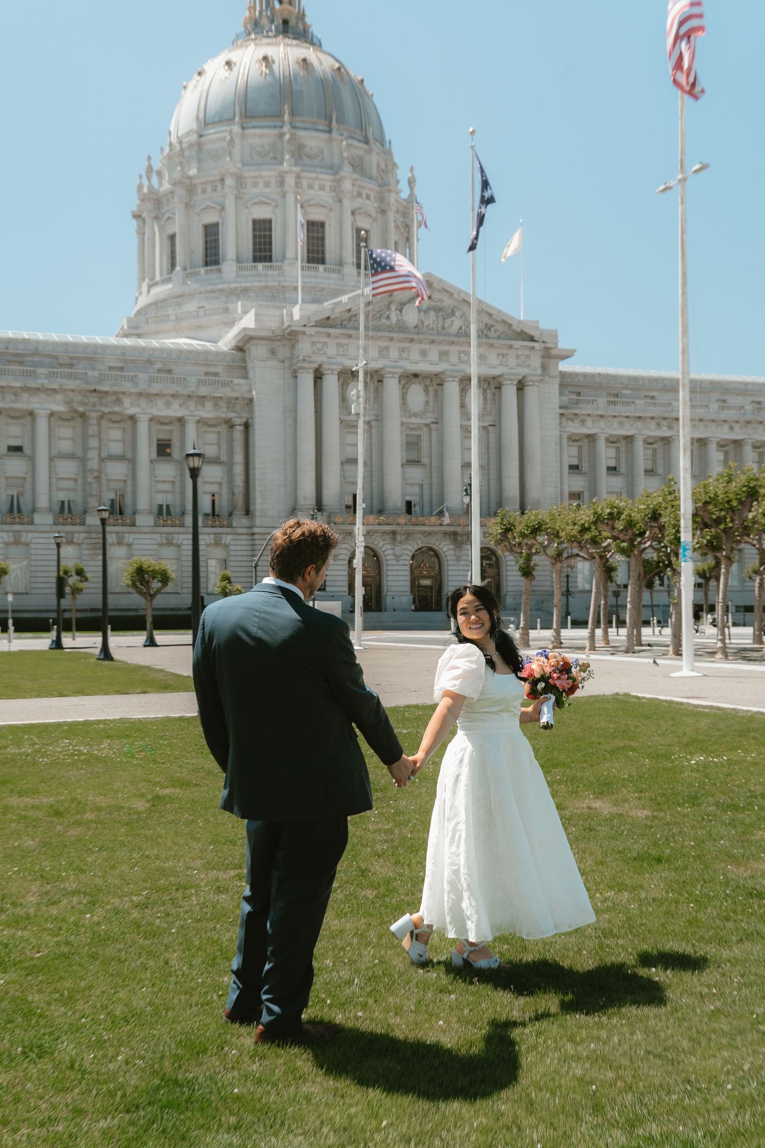 Bride and groom hold hands on lawn in front of San Francisco City Hall on wedding day.