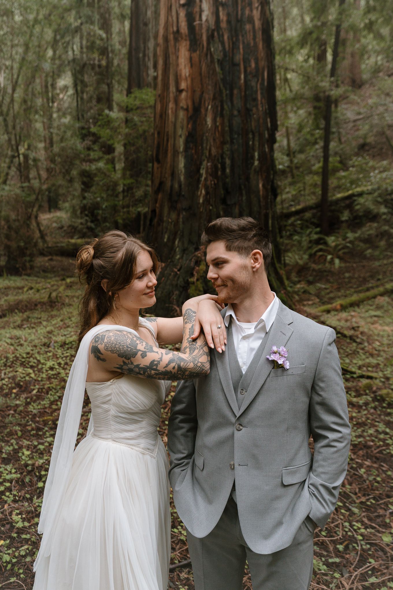 Bride with tattoos in white gown and groom in gray suit share a romantic moment in a redwood forest.