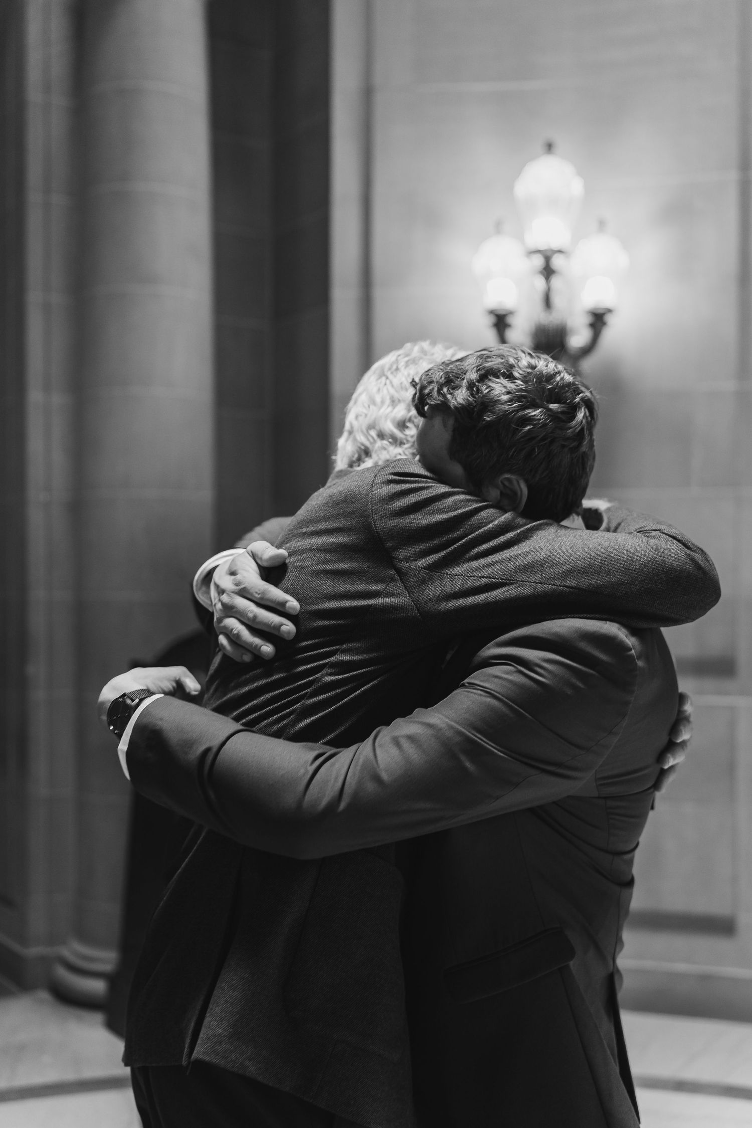 Two grooms share an emotional embrace in a dimly lit elegant hall on their wedding day.