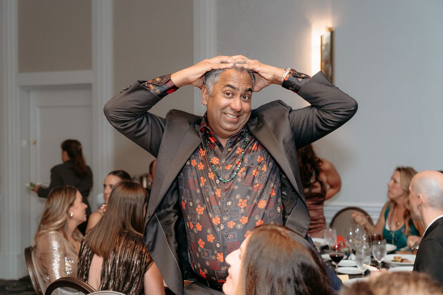 Man in floral shirt and blazer gestures expressively at an elegant indoor dinner event.