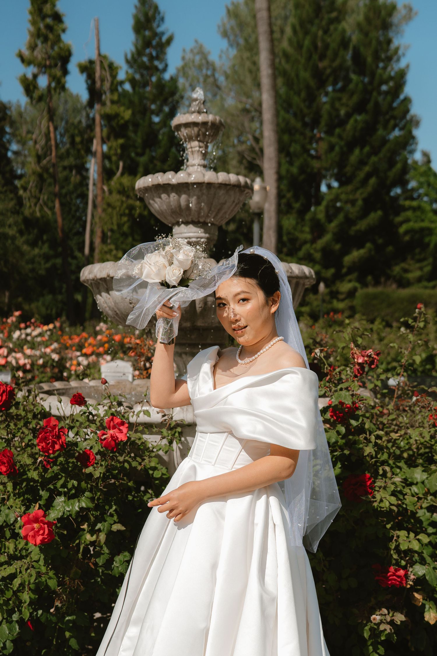 Asian bride in white gown and veil poses with bouquet near stone fountain surrounded by red roses in garden.