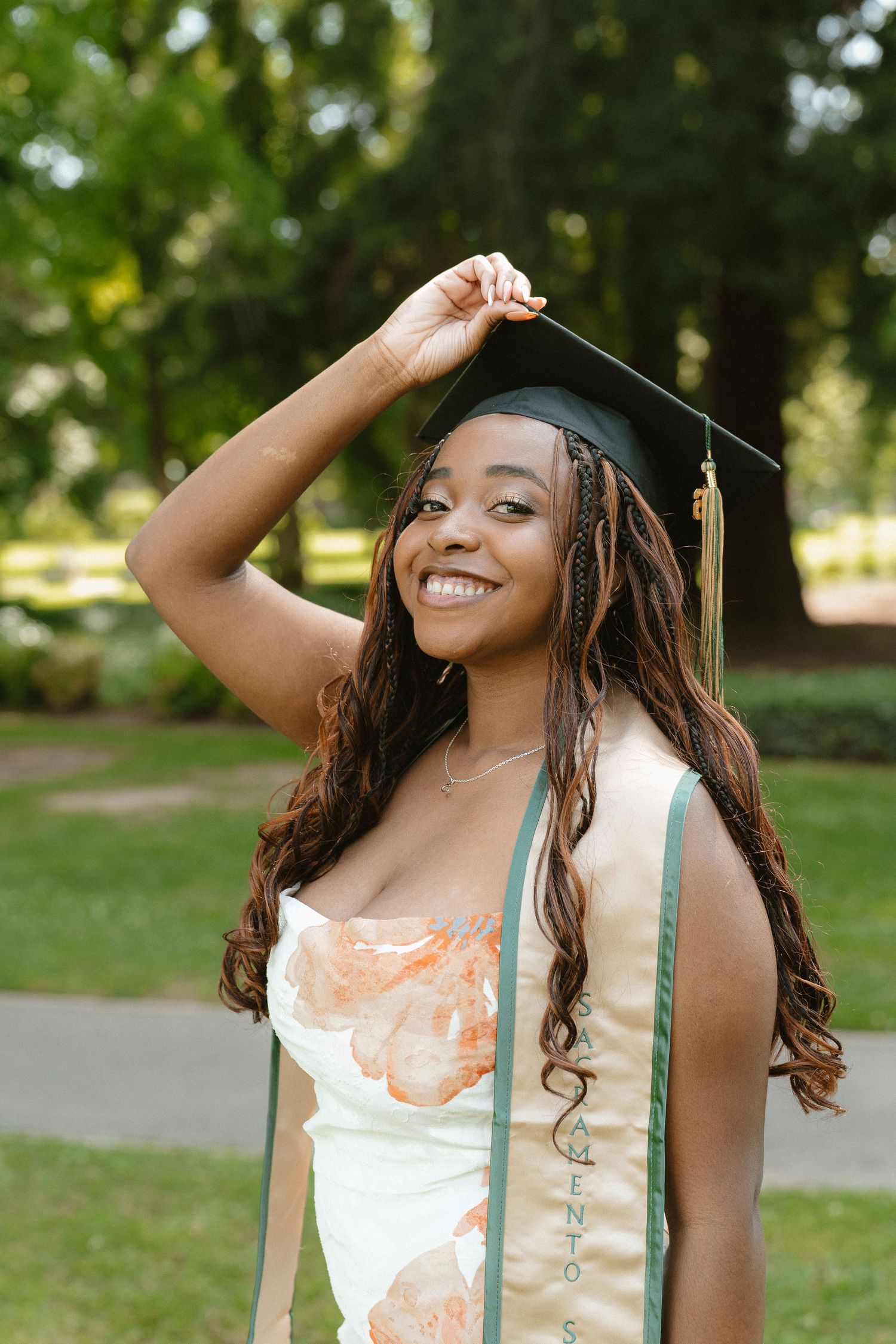 Smiling young woman in graduation cap and green sash poses outdoors in a floral dress on her graduation day.