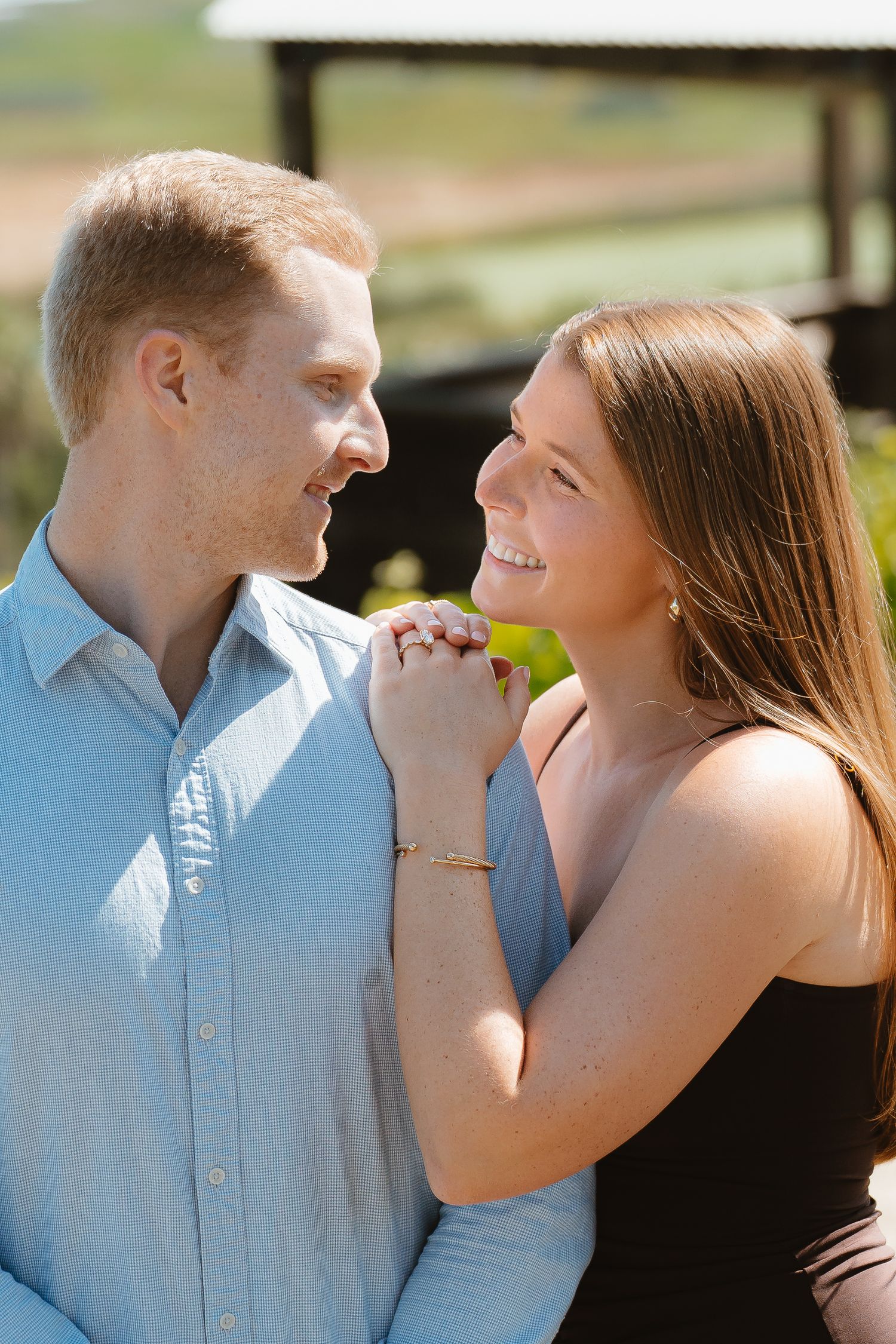 A smiling couple holds hands outdoors, gazing at each other affectionately in warm sunlight near a rustic barn.