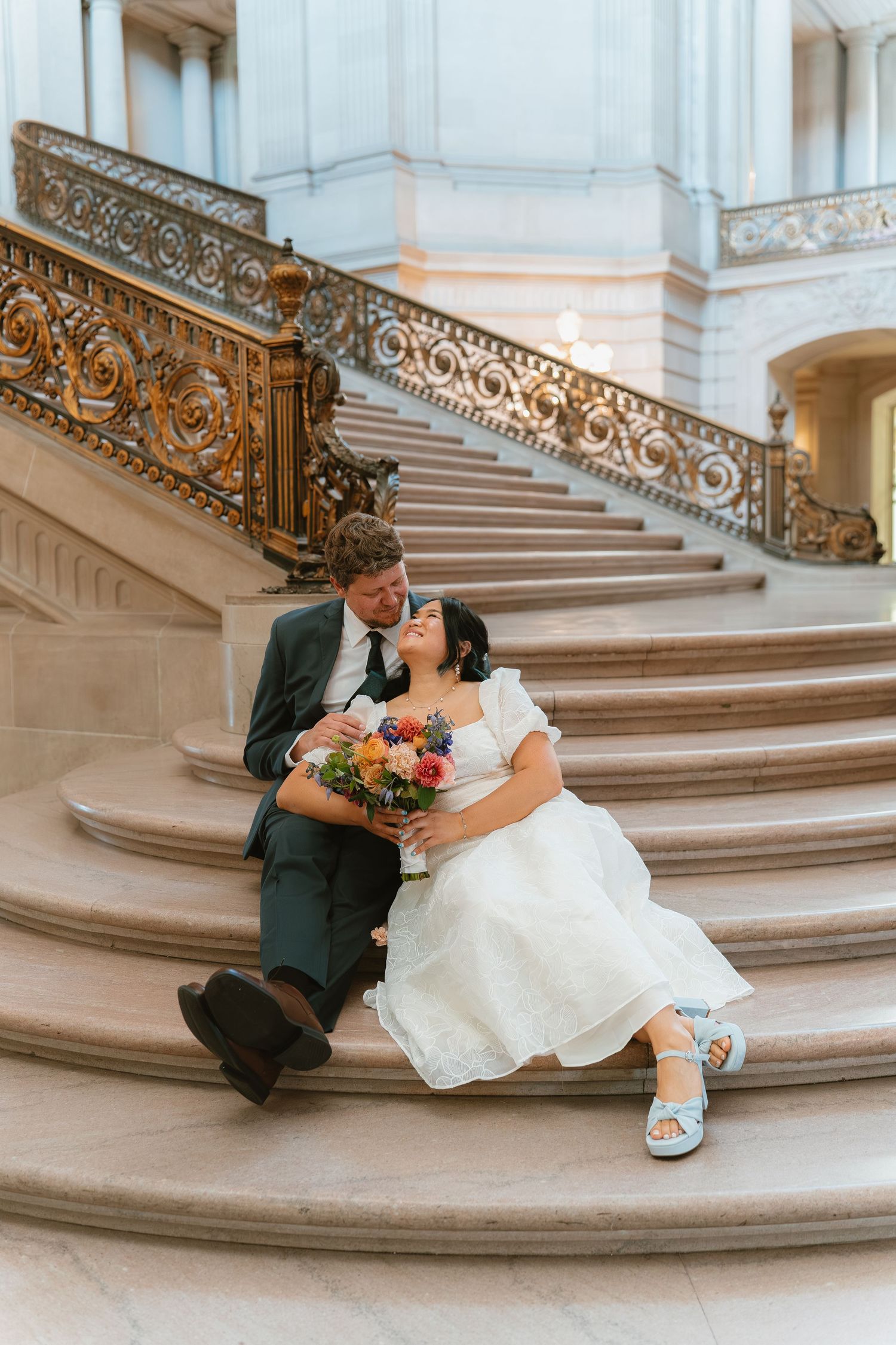 Bride and groom sit closely on grand marble staircase at San Francisco City Hall wedding.