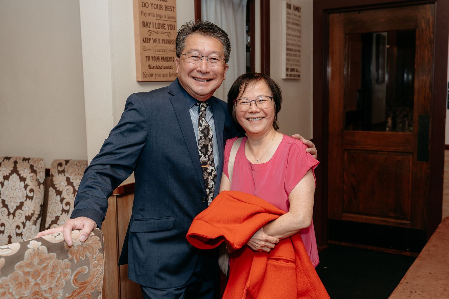 An older Asian couple smiling and posing together at an indoor event, woman in pink holding an orange jacket.