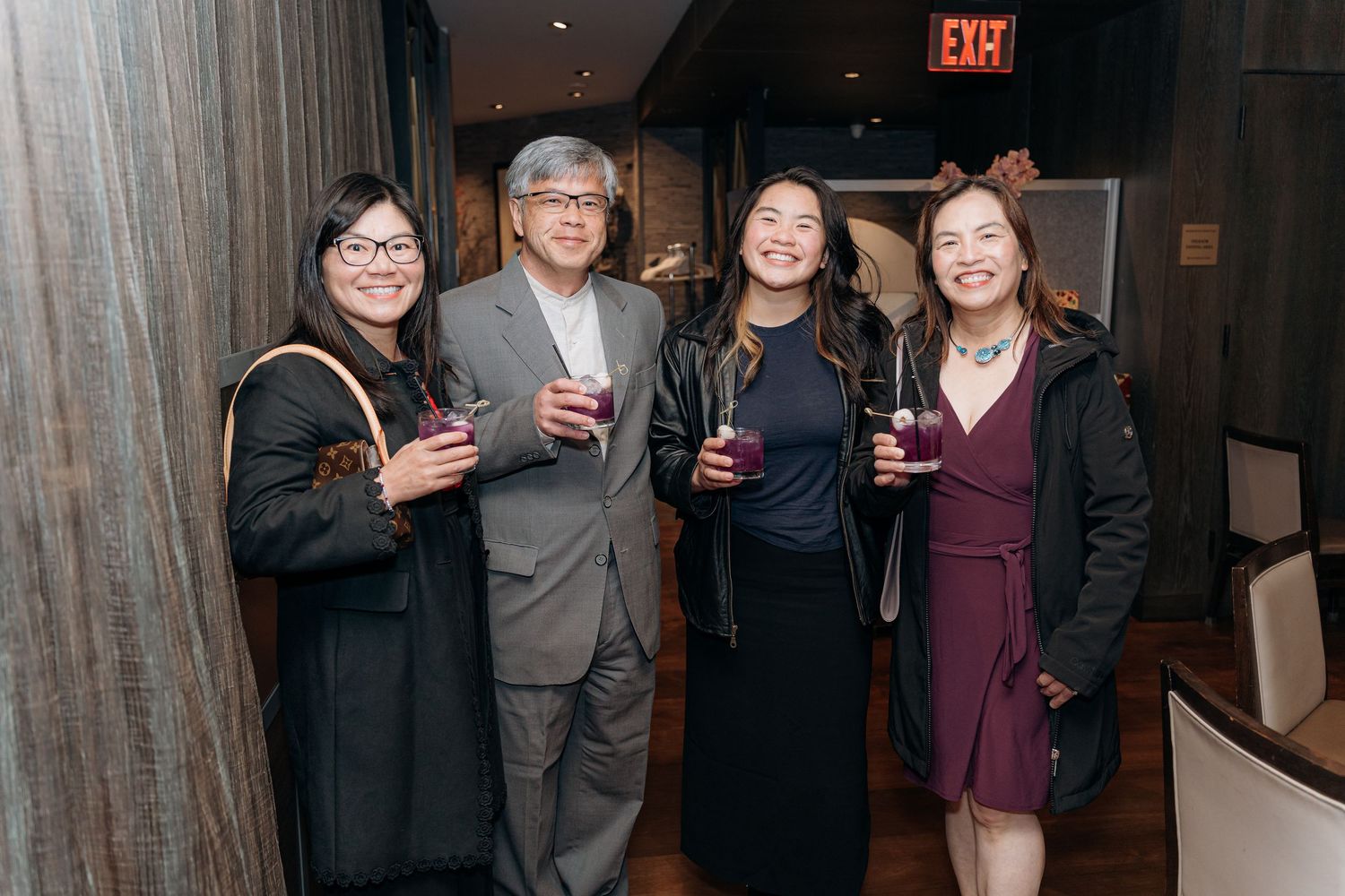 Four guests smile and hold drinks at an indoor evening social event near a warmly lit bar area.