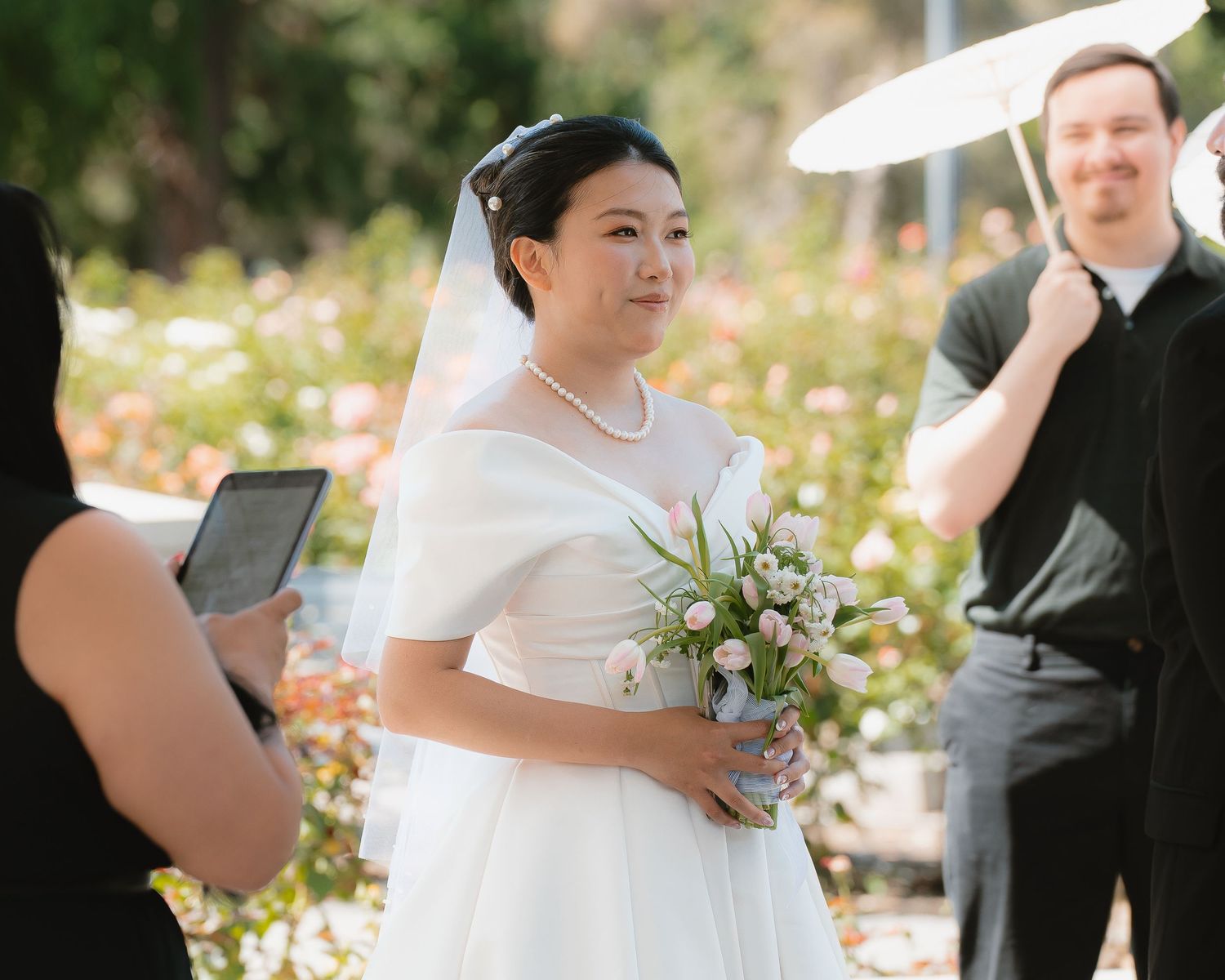 Bride in white gown and veil holds bouquet, smiling during outdoor wedding ceremony with floral backdrop.