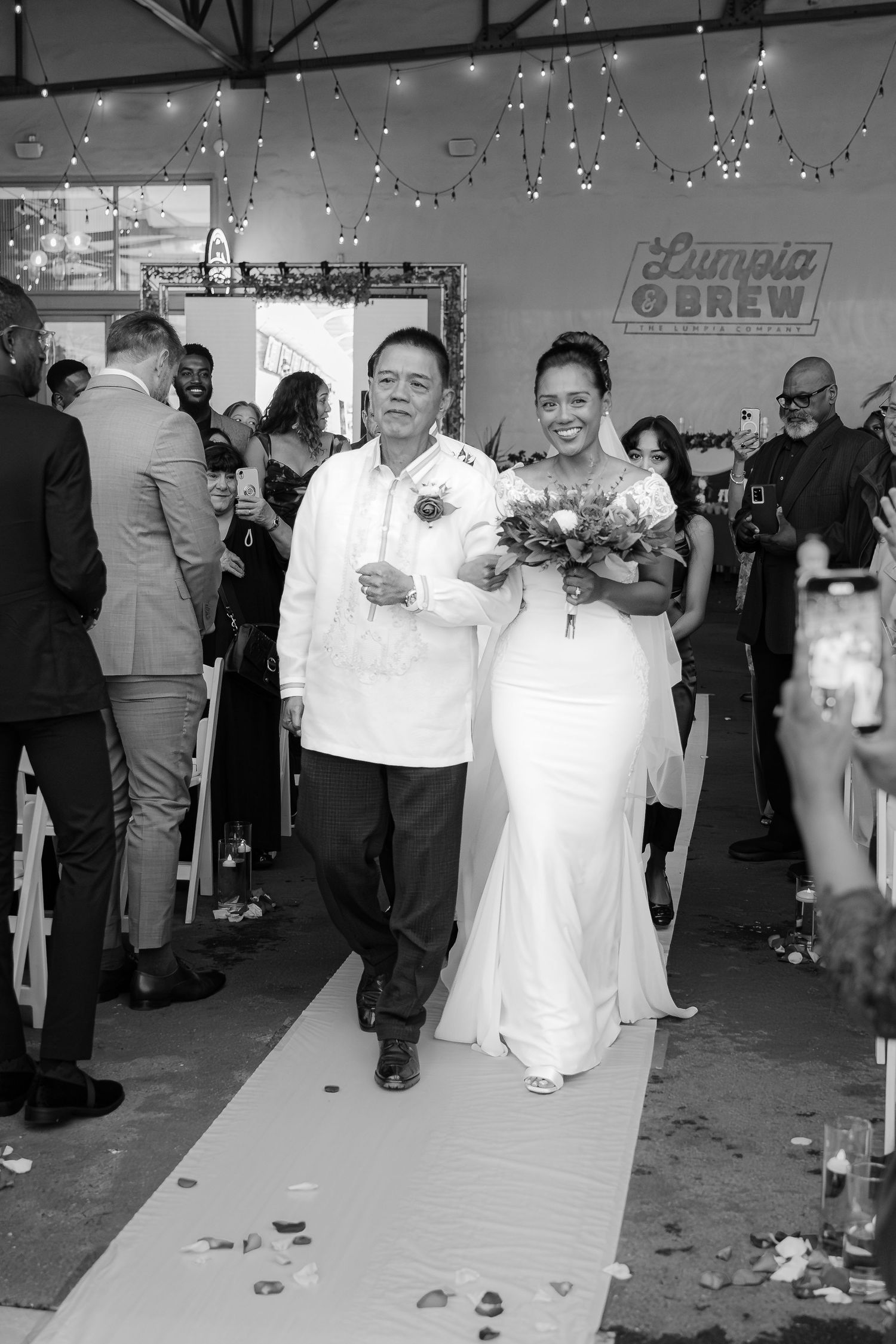 Black and white photo of a bride walking down the aisle with her father at a Lumpia and Brew wedding ceremony.
