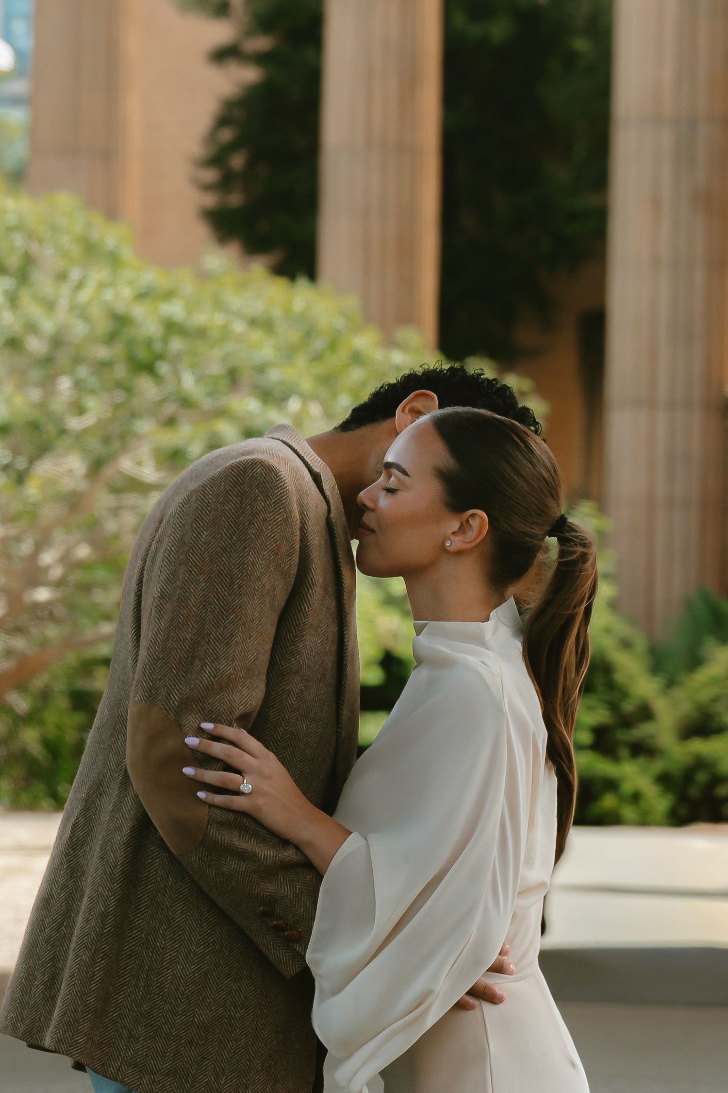 Couple sharing a romantic kiss outdoors, woman in white blouse with engagement ring, man in brown blazer, greenery background.