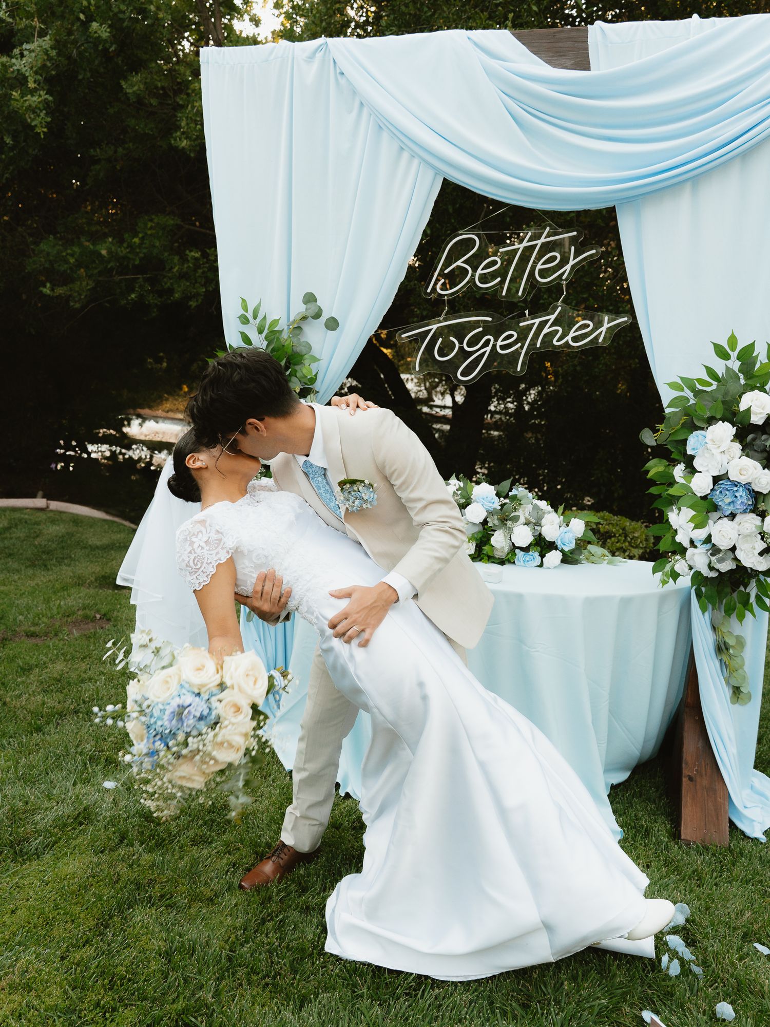 A bride and groom share a romantic dip kiss under a blue draped arch with a neon Better Together sign.