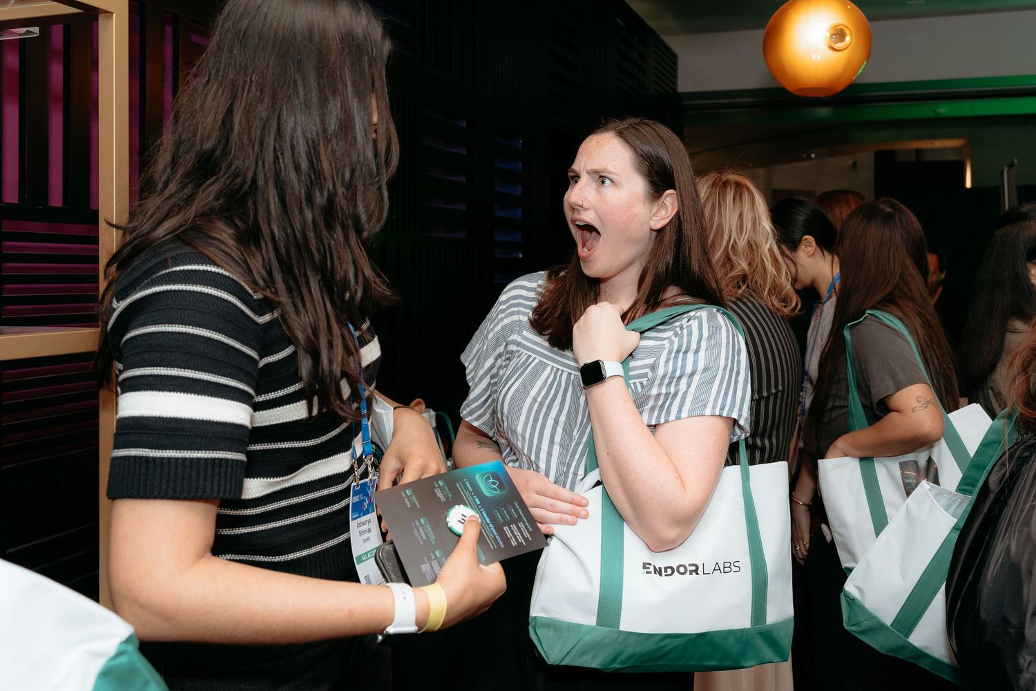 corporate event for tech company in Two women networking at an event, one reacting with surprise, holding a tote bag at an indoor gathering.