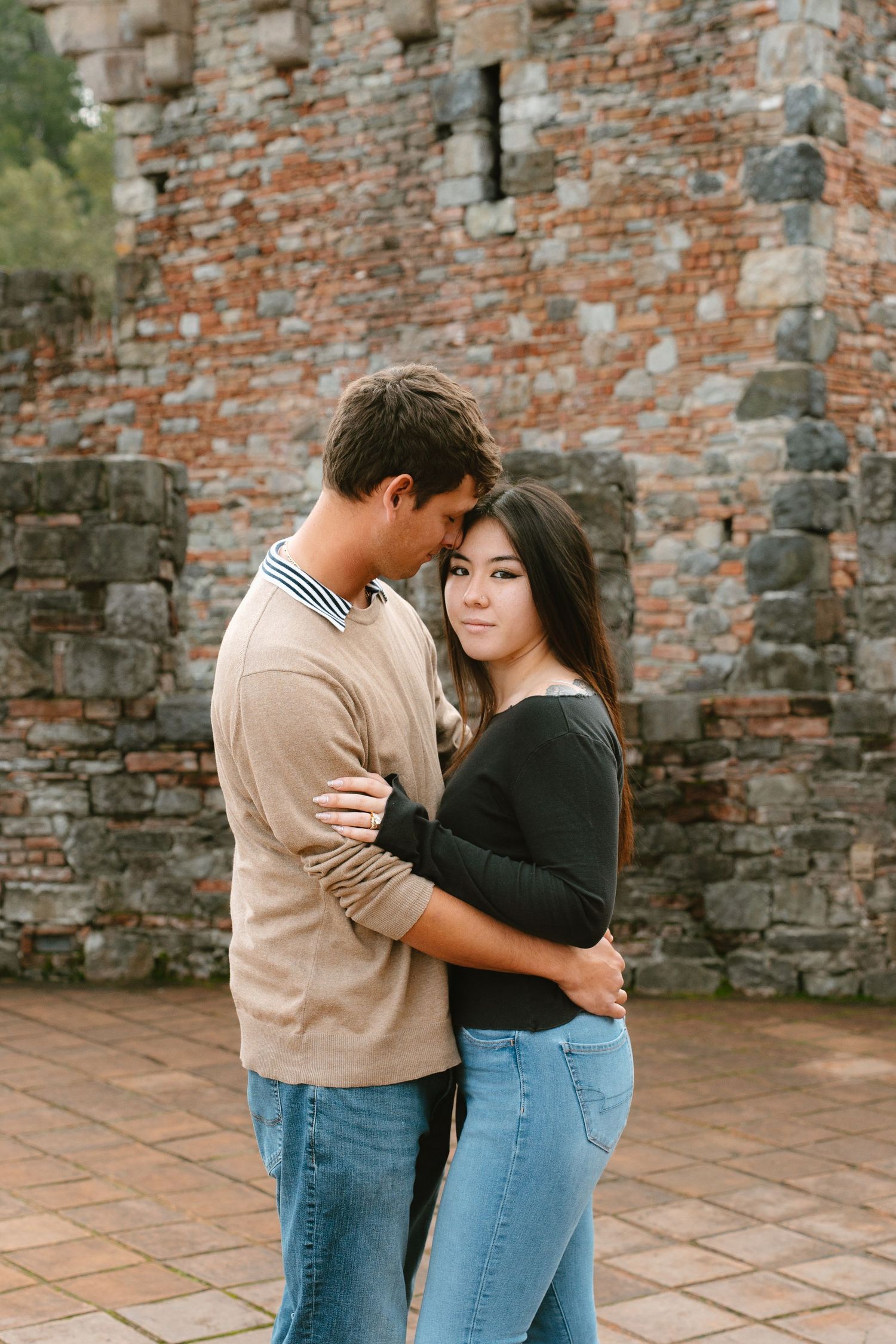 A romantic couple embraces against a rustic brick wall backdrop while wearing casual denim and neutral tones in Napa Valley winery.