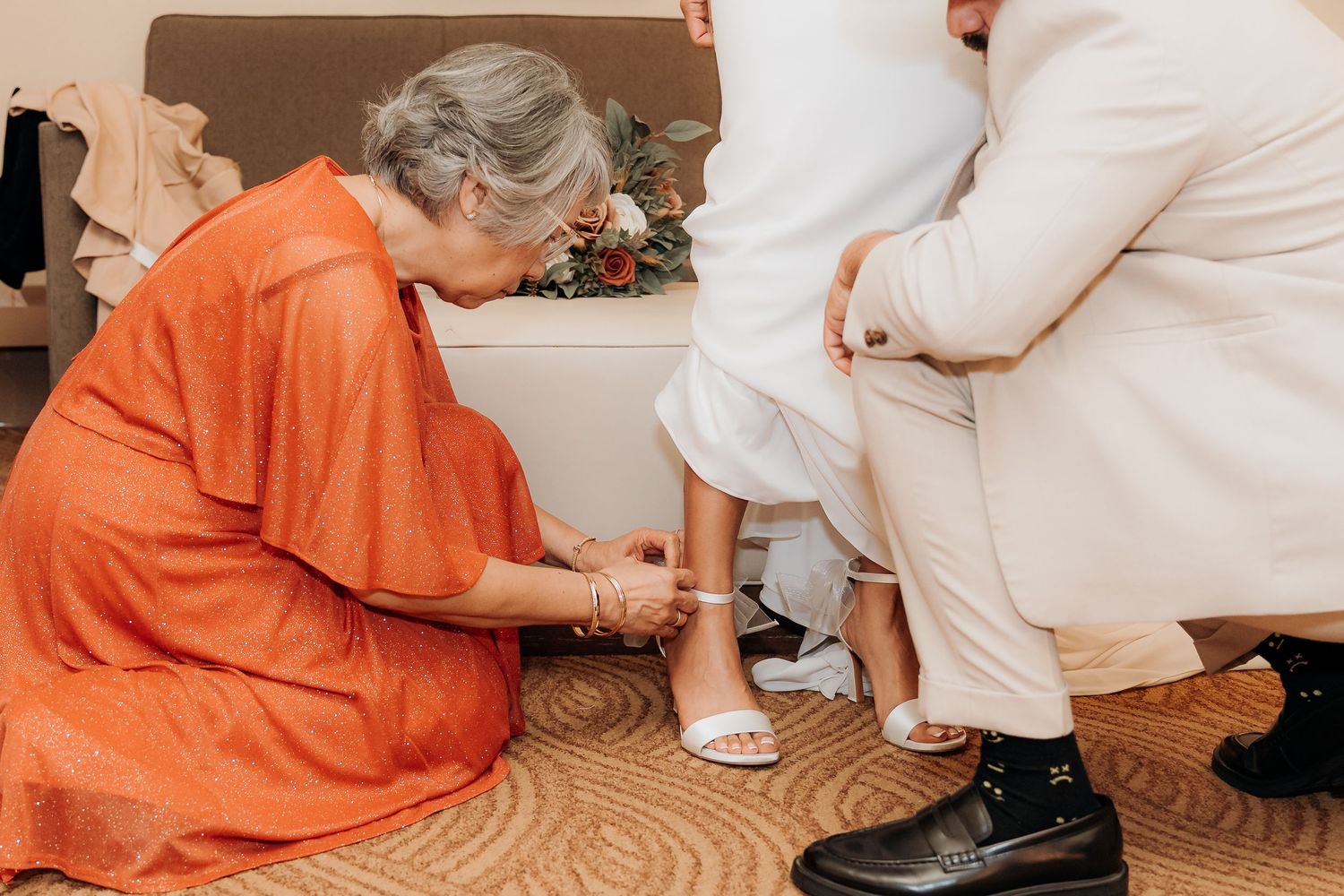 Elderly woman in orange dress kneeling to help bride with white wedding shoes during bridal preparation.